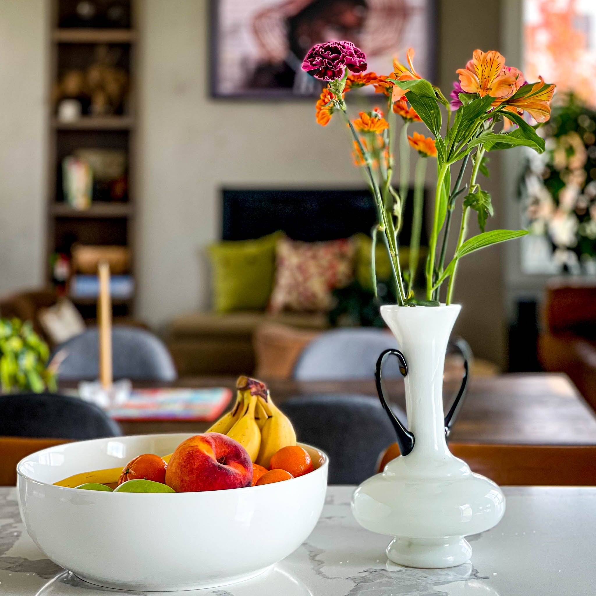 White bowl of bananas, peaches, oranges, and limes beside a white vase of orange alstroemeria and magenta carnations on a marble table, a colorful photo ready to inspire fiber art color choices