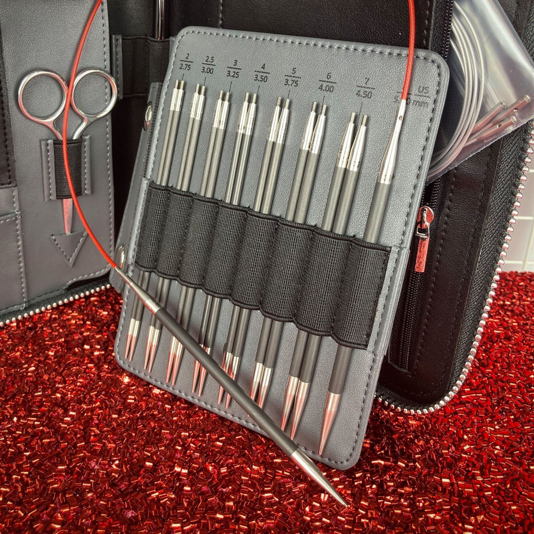 ChiaoGoo FORTÉ 2.0 interchangeable tips US-2 through US-8 in a gray fold-out case with a red TWIST cable threaded through, embroidery scissors and the zippered case behind, on a red beaded background
