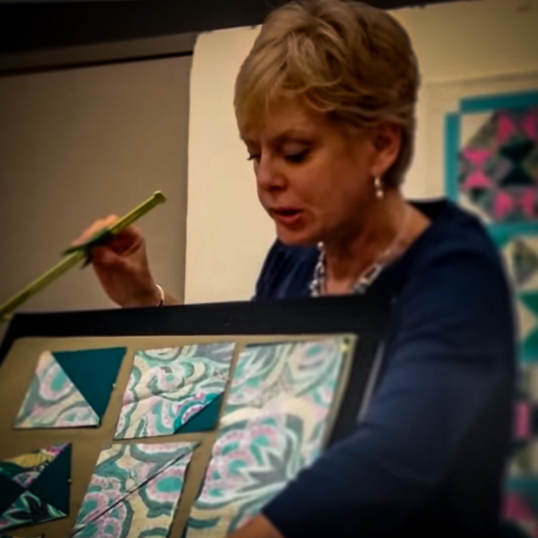 Nancy Zieman pointing to a demonstration board of teal and floral half-square triangle quilt blocks, with a pieced quilt on the wall behind her