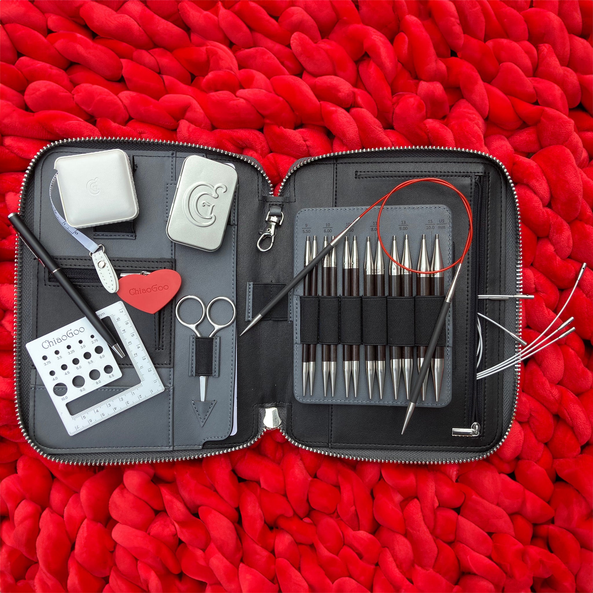 ChiaoGoo FORTÉ 2.0 Interchangeable Set open in its charcoal zippered case on a red chunky knit blanket, showing blackwood and carbon fiber tips, a red TWIST cable, needle gauge, scissors, and silver accessory tins