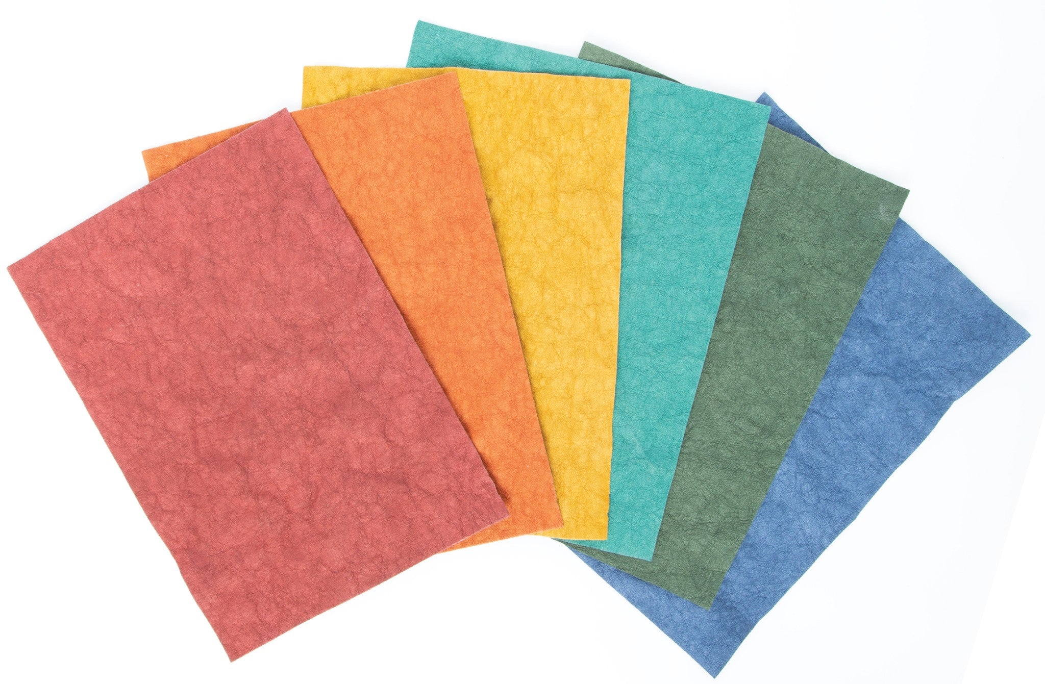 Six sheets of Kraft-Tex Designer hand-dyed prewashed paper fabric fanned out in Bordeaux, Terra Cotta, Ochre, Teal, Alpine Green, and Atlantic Blue, showing the soft crinkled leather-like surface.