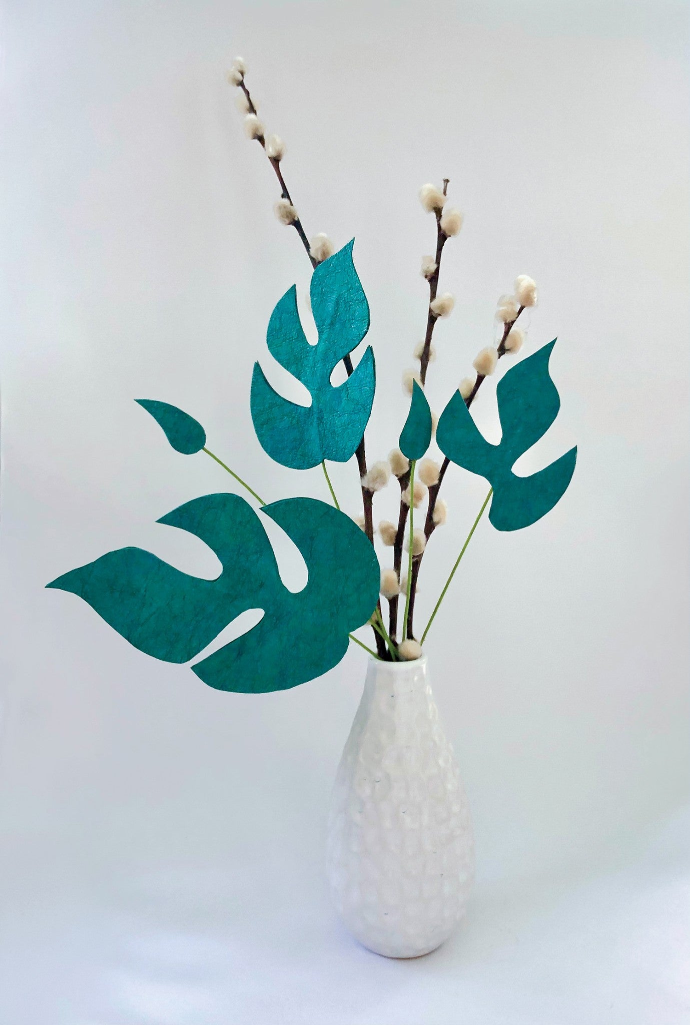Decorative monstera-style leaves cut from Teal Kraft-Tex Designer paper fabric, arranged on stems with pussy willow branches in a white vase.