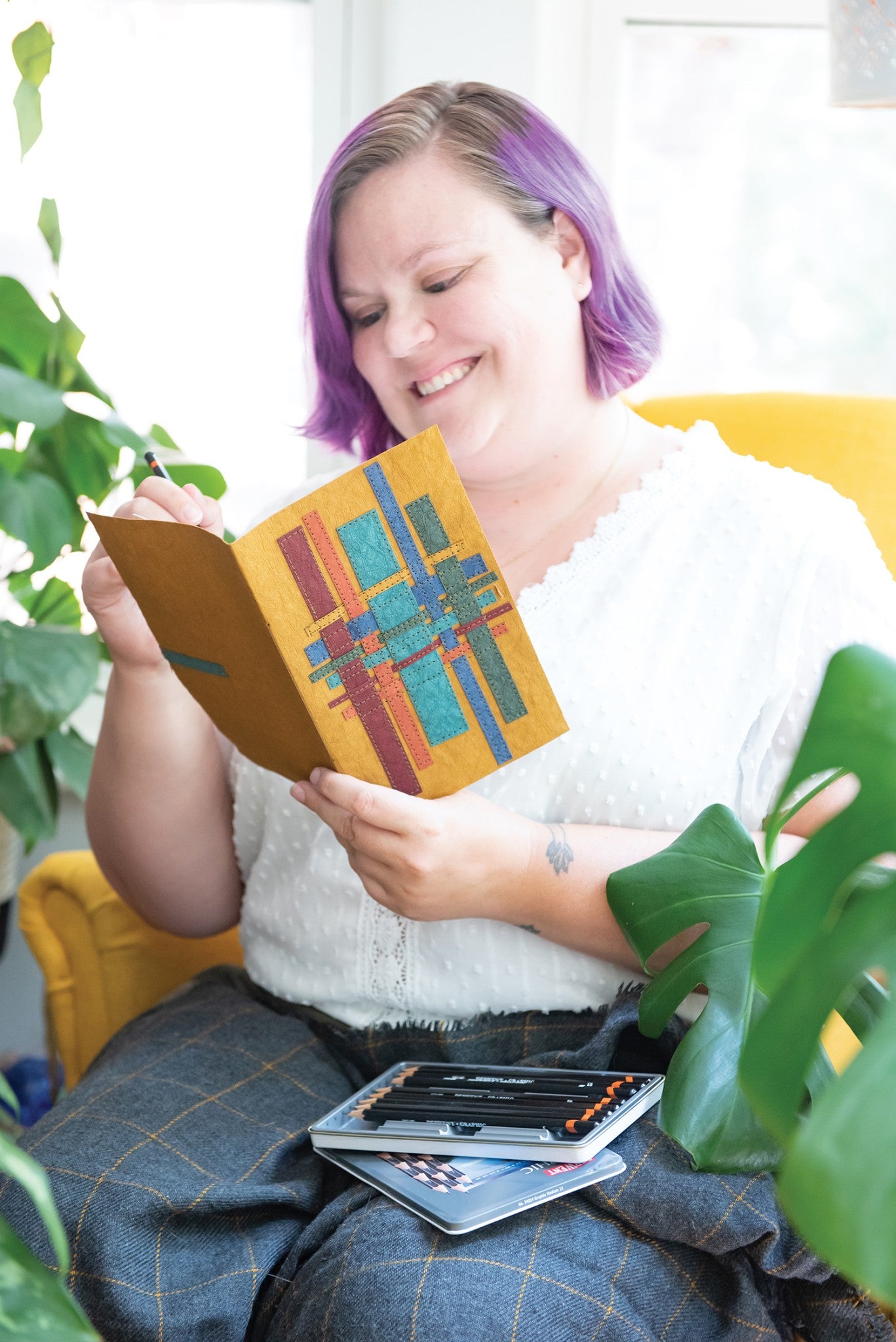 A person looking at an Ochre Kraft-Tex Designer paper fabric journal cover woven with strips in the sampler colors, with colored pencils nearby, by C&T Publishing.