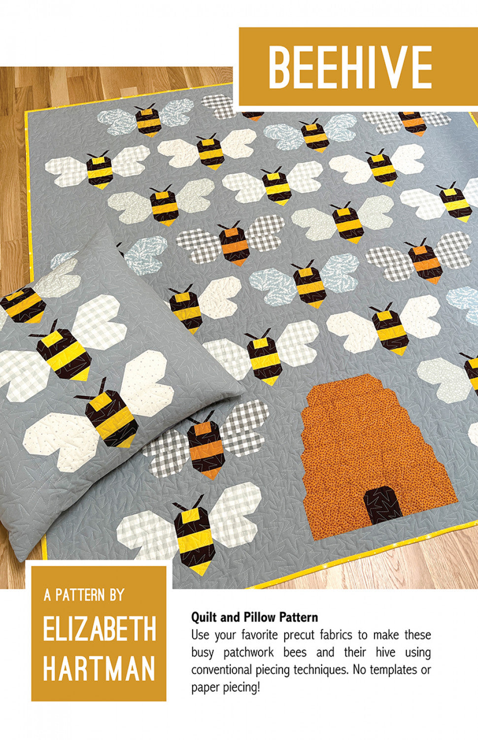 Beehive quilt pattern cover by Elizabeth Hartman. Large quilt and pillow displayed on a wood floor show patchwork bumblebees with gold and brown striped bodies and varied neutral wings including white, gingham, and subtle prints on a gray background, with a warm orange honey hive block in the lower right corner. Yellow binding visible.