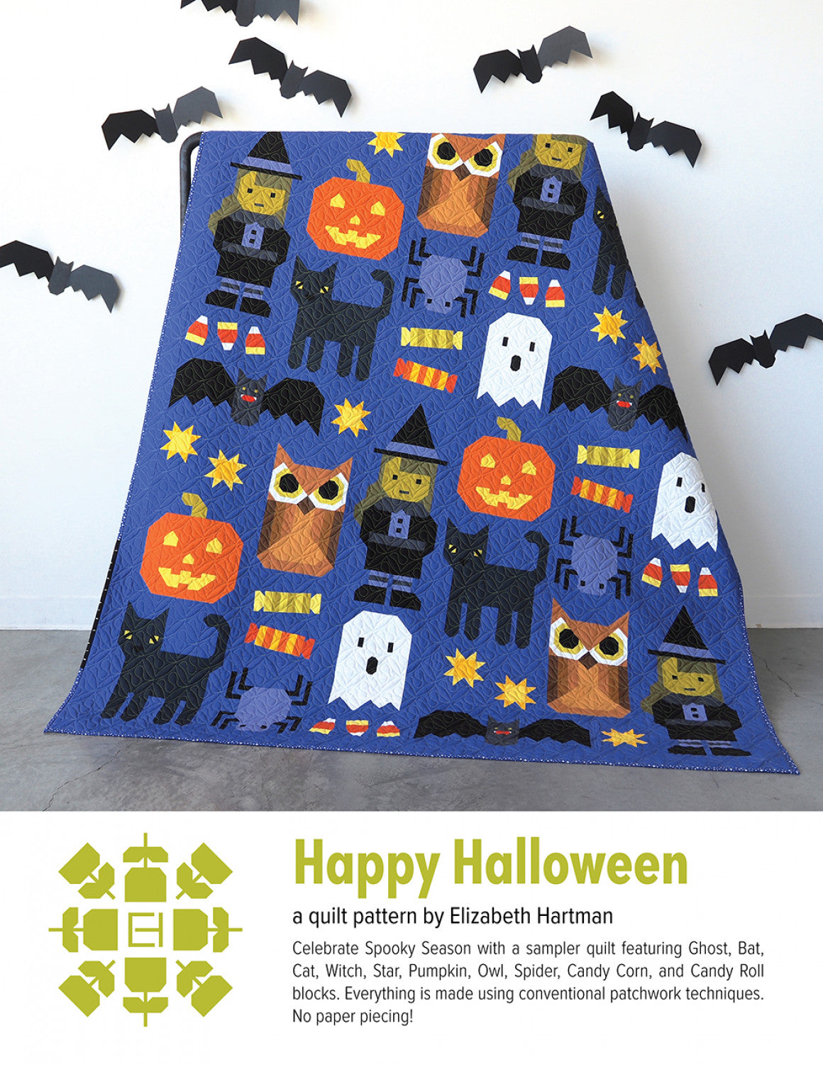 Happy Halloween quilt pattern cover by Elizabeth Hartman. Large 66 by 82 inch quilt leaning against a wall with paper bat decorations shows patchwork Halloween blocks including Ghost, Bat, Black Cat, Witch, Star, Jack-o-Lantern Pumpkin, Owl, Spider, Candy Corn, and Candy Roll on a rich cobalt blue-purple background.