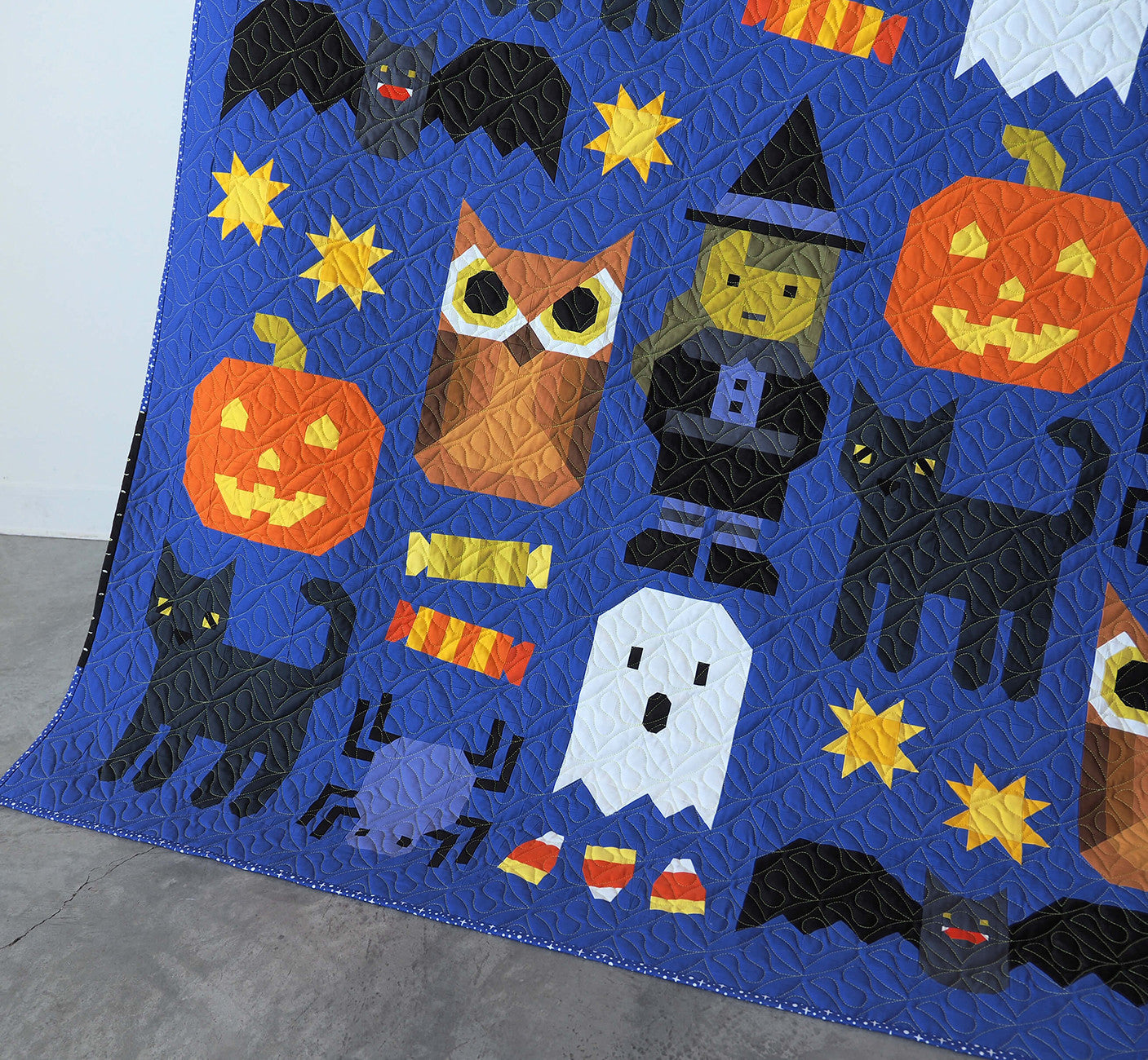 Detail of lower section of the Happy Halloween large quilt by Elizabeth Hartman showing patchwork blocks including black Bats with wings spread, yellow Stars, an Owl, a Witch, an orange Pumpkin, a white Ghost, black Cats, Candy Corn, and Candy Roll blocks on cobalt blue-purple background.