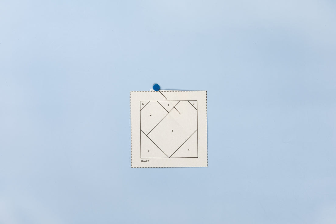 A foundation pattern printed on Carol Doak's Foundation Paper from C&T Publishing, pinned to a pale blue background. The sheet shows a heart block drafted in fine black lines with sections numbered one through seven, a dashed cutting border, and the label Heart 2 in the lower corner.