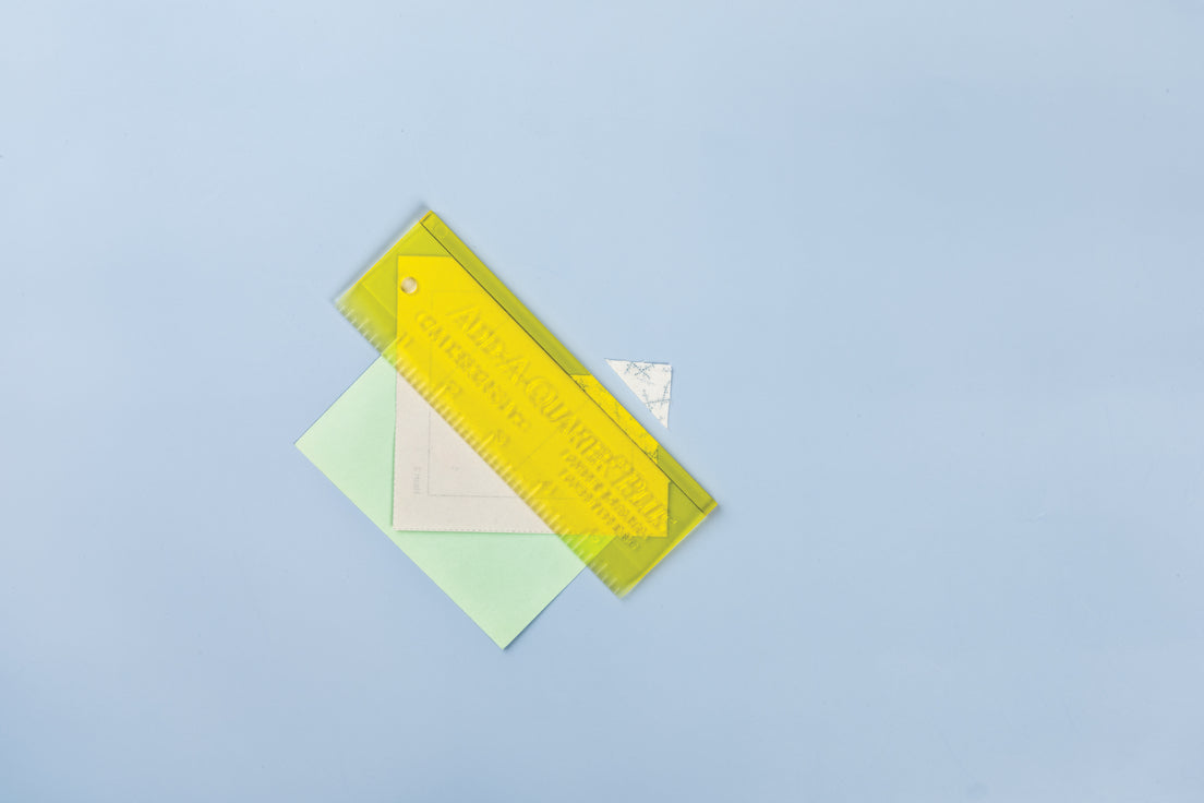 Carol Doak's Foundation Paper from C&T Publishing folded back along a stitching line, with a yellow Add-A-Quarter ruler laid against the fold to trim the seam allowance to a quarter inch. A pale green card supports the fold. The ruler and card are not included with the paper.
