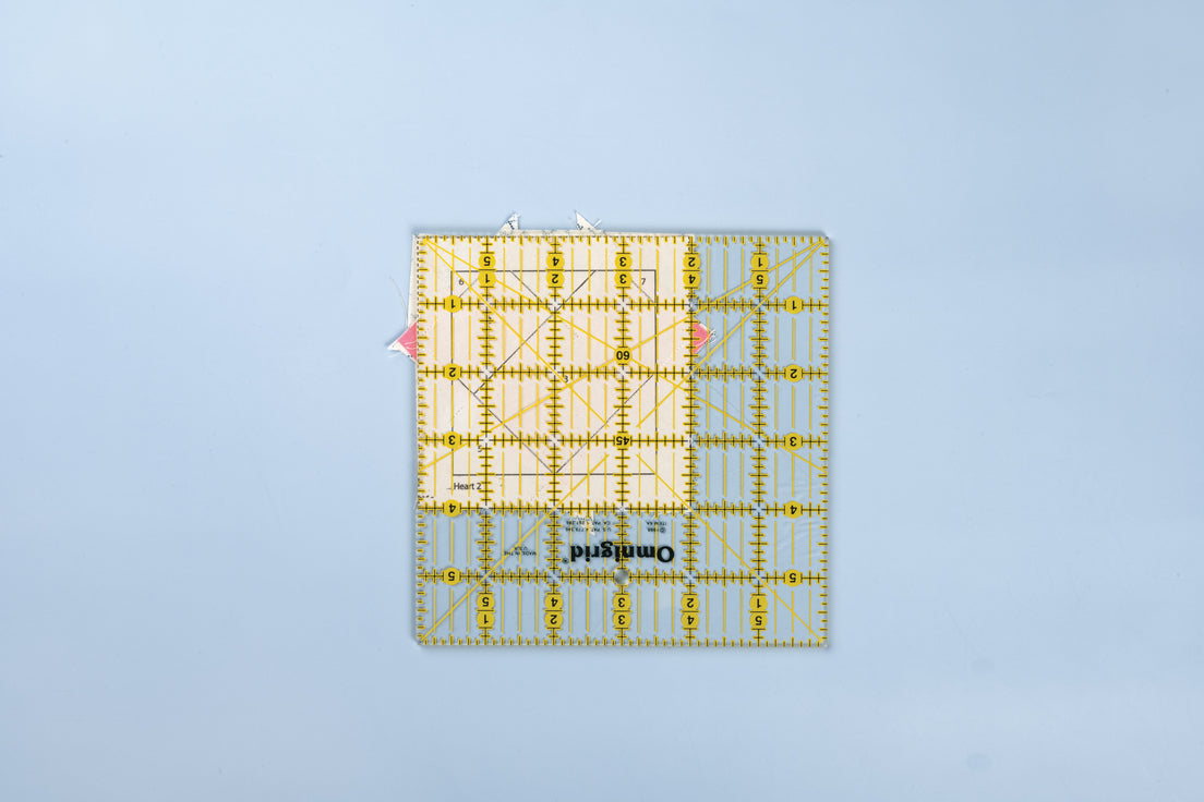 A pieced heart block still attached to Carol Doak's Foundation Paper from C&T Publishing, squared up under a yellow Omnigrid square ruler on a pale blue background. Trimmed points and threads show along the edges. The ruler is not included with the paper.