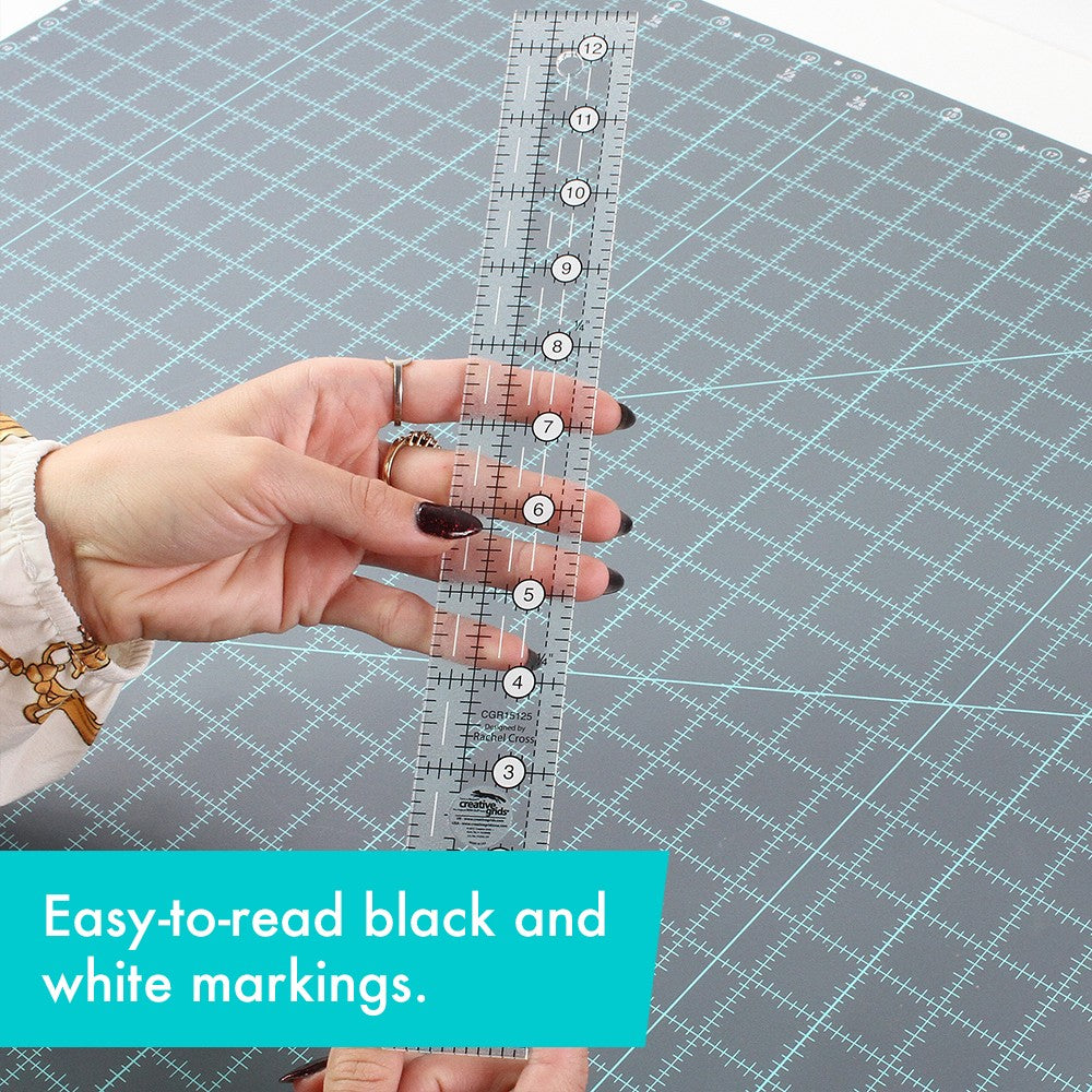 Hand holding the Creative Grids 1-1/2 x 12-1/2 inch ruler CGR15125 upright over a gray cutting mat, with a brand caption about the black and white markings.