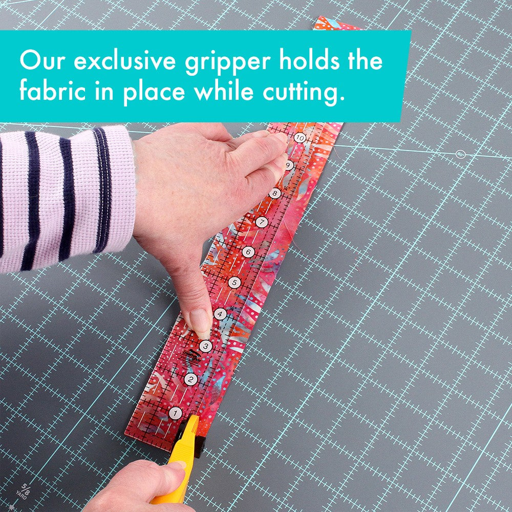 Hands pressing the Creative Grids 1-1/2 x 12-1/2 inch ruler CGR15125 on red batik fabric while a rotary cutter trims beside it, with a brand caption about the gripper holding fabric in place.