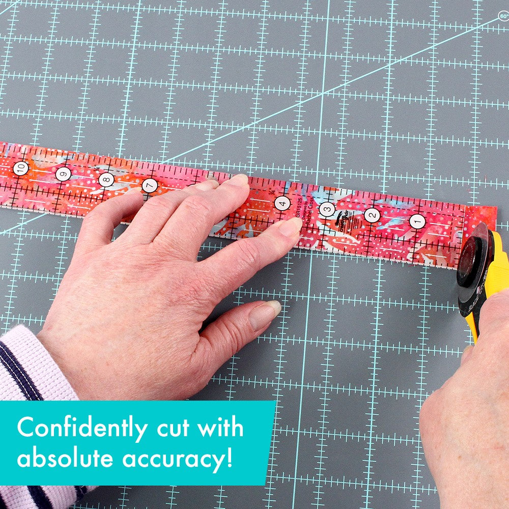 Hand holding the Creative Grids 1-1/2 x 12-1/2 inch ruler CGR15125 flat on red batik fabric with a rotary cutter, with a brand caption about cutting with accuracy.