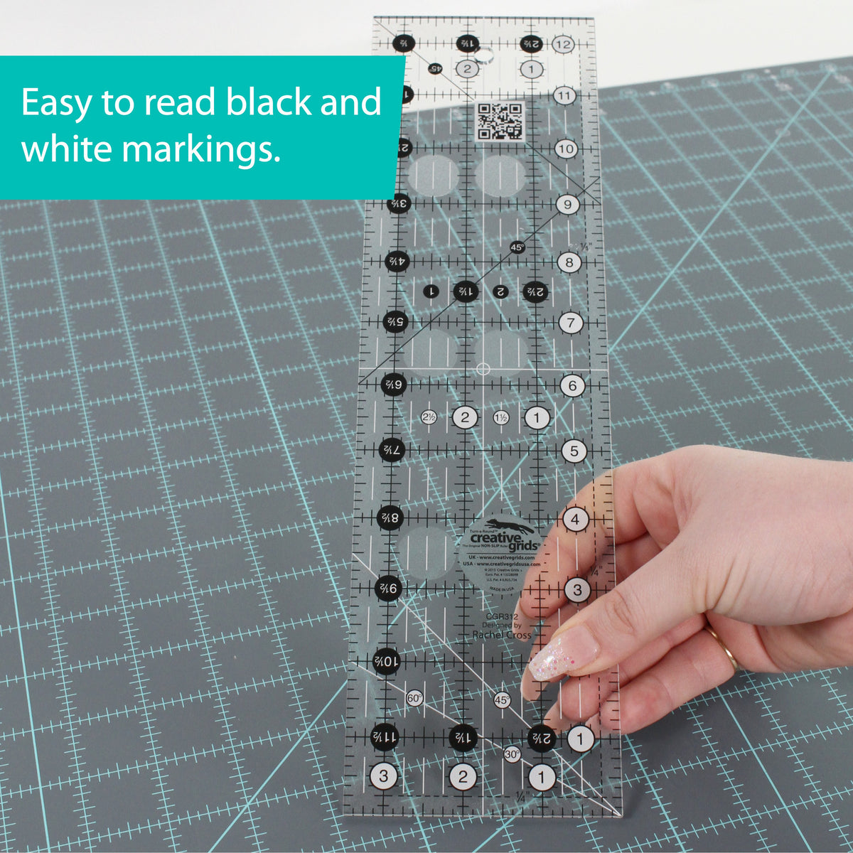 Hand holding the Creative Grids 3-1/2 x 12-1/2 inch ruler CGR312 upright over a gray cutting mat, with a brand caption about the black and white markings.