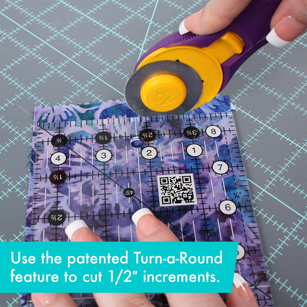 Creative Grids 4-1/2 x 8-1/2 inch ruler CGR48 on purple batik fabric with a rotary cutter at the top edge, with a brand caption about using the Turn-a-Round feature to cut half-inch increments.
