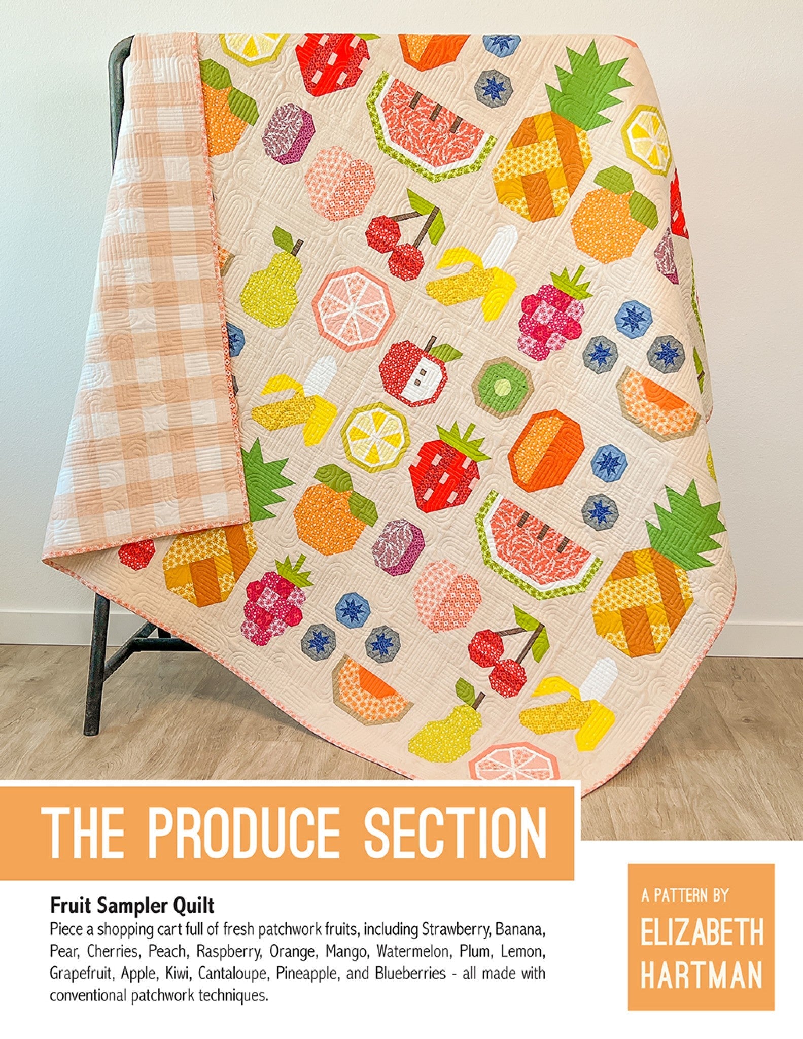 The Produce Section quilt pattern cover by Elizabeth Hartman. Large 75 by 75 inch quilt propped on a stand shows 17 different patchwork fruit blocks including strawberry, banana, pear, cherries, peach, raspberry, orange, mango, watermelon, plum, lemon, grapefruit, apple, kiwi, cantaloupe, pineapple, and blueberries scattered across a warm cream background. The pink gingham backing is visible.
