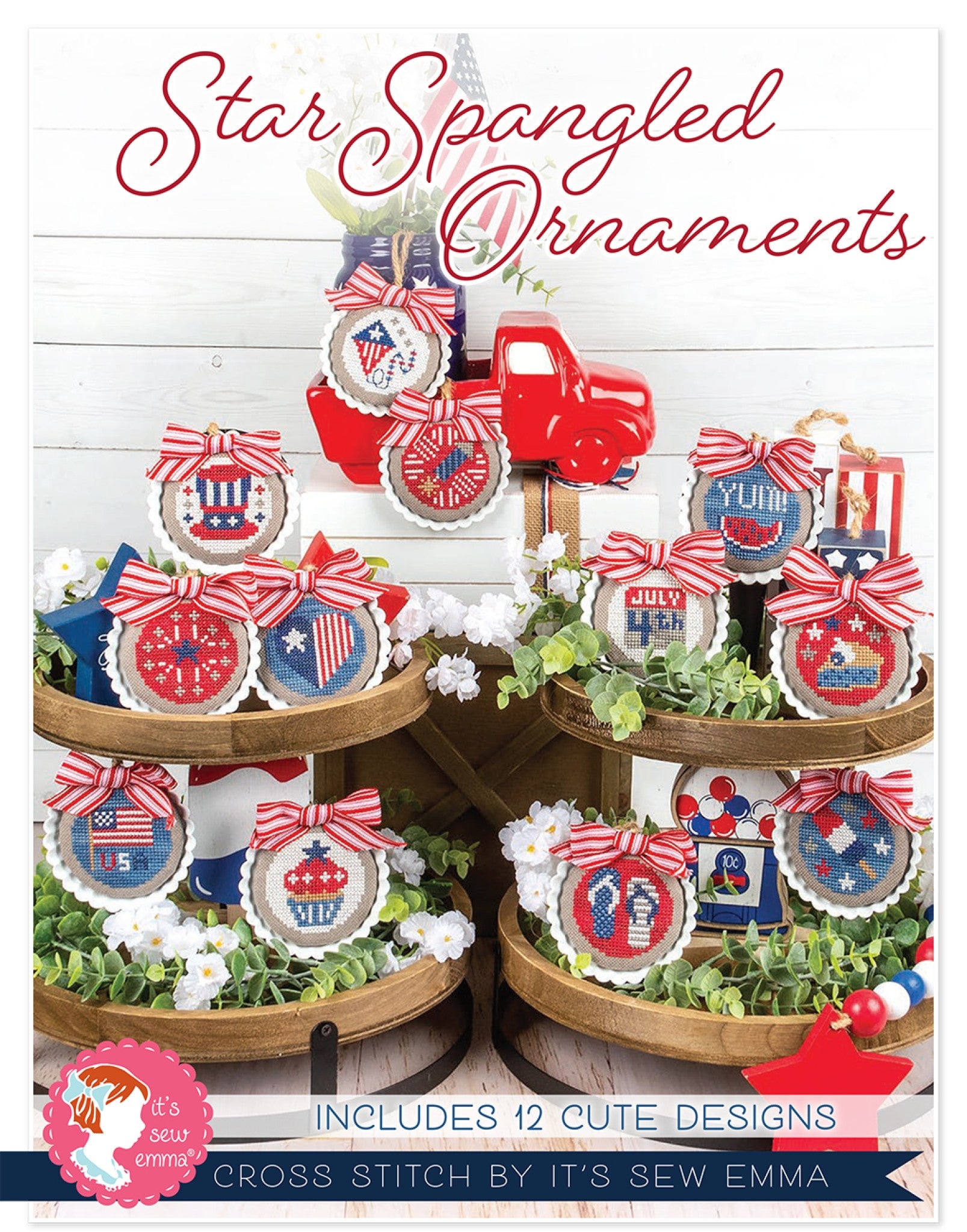 Front cover of Star Spangled Ornaments cross stitch pattern by It's Sew Emma. A tiered wooden tray displays a dozen round red, white and blue cross stitch ornaments tied with red-and-white striped bows, including Uncle Sam's hat, a flag, a watermelon slice reading YUM, a cupcake, flip flops, a gumball machine, fireworks and a JULY 4TH banner, on pebble-tan cloth styled with greenery and white flowers. Text reads Includes 12 Cute Designs.