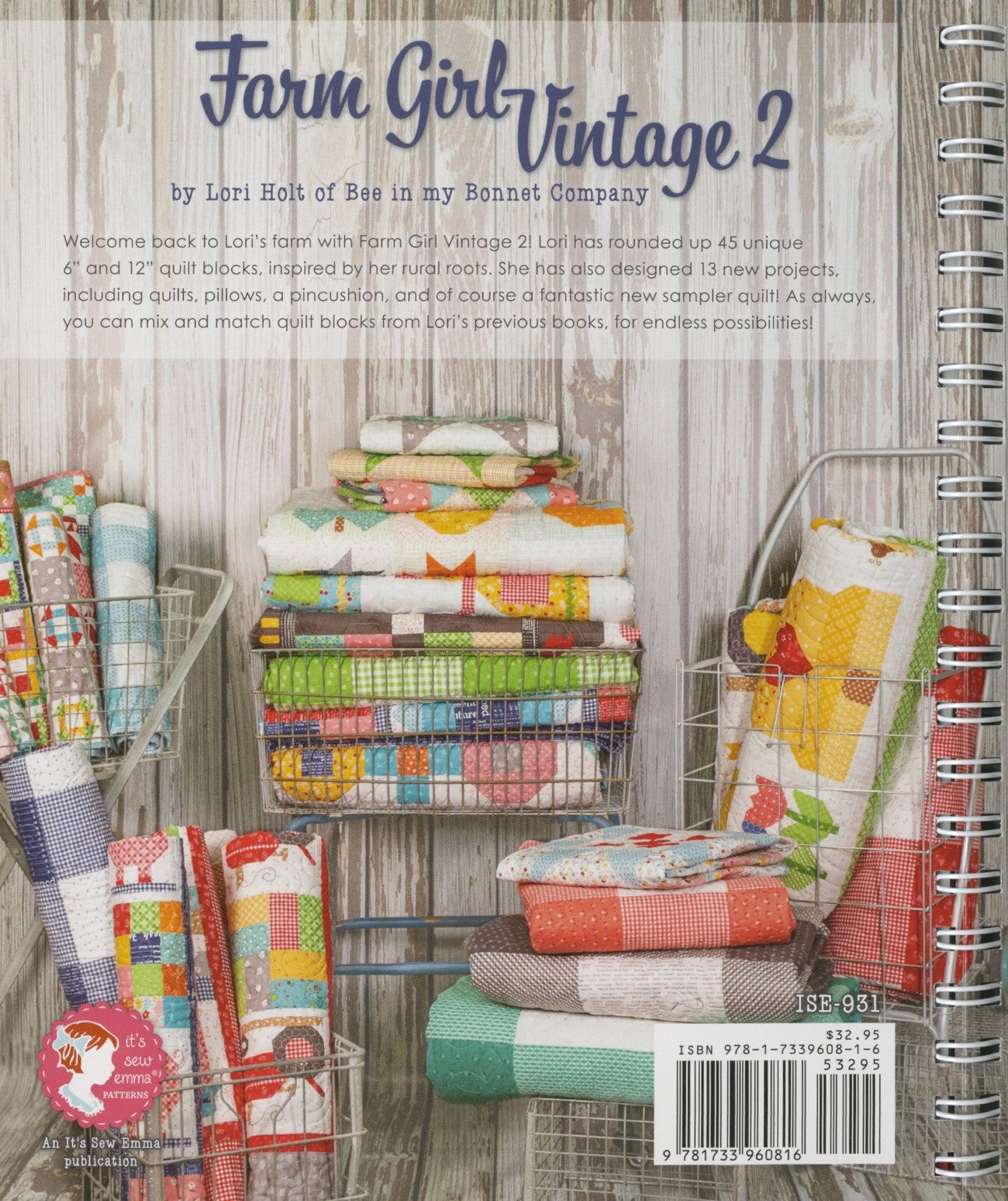 Back cover of Farm Girl Vintage 2 by Lori Holt for It's Sew Emma. Folded scrappy patchwork quilts fill wire baskets and a vintage cart against weathered wood, with the It's Sew Emma logo and item number ISE-931.