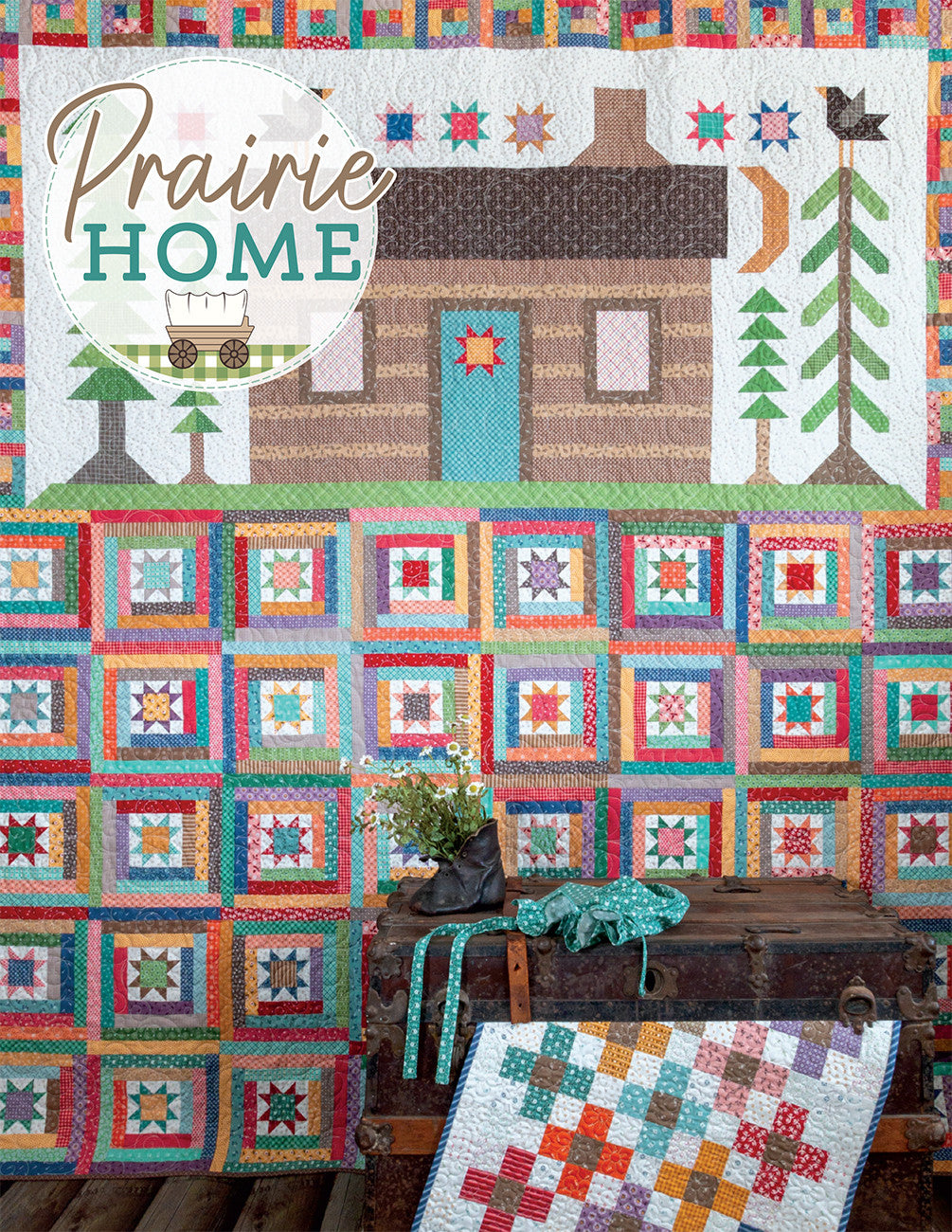The Prairie Home sampler quilt by Lori Holt for It's Sew Emma, a log cabin and pieced star design, displayed on a wall above a wooden trunk styled with flowers, with a coordinating scrappy quilt draped nearby.