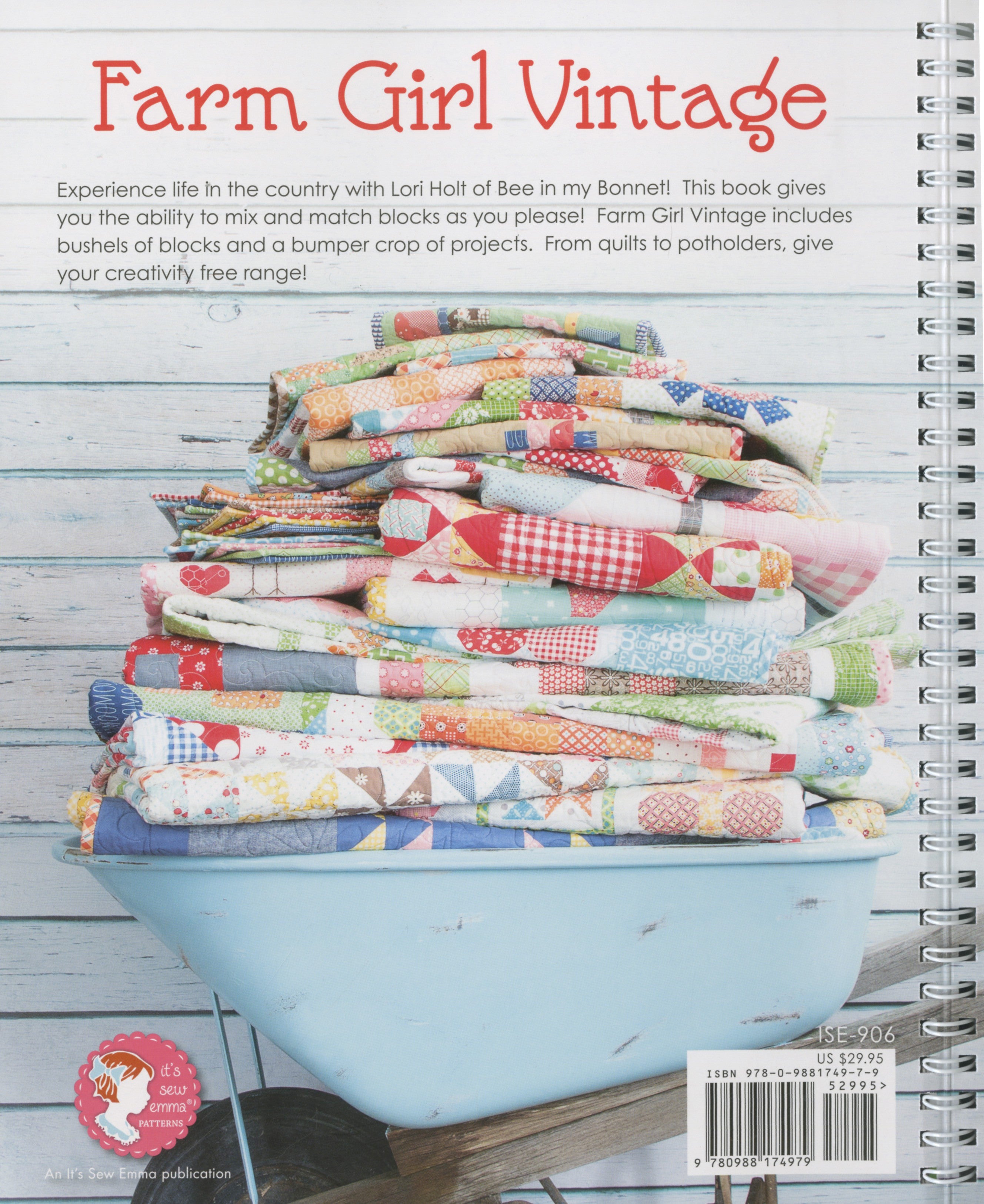 Back cover of Farm Girl Vintage by Lori Holt for It's Sew Emma. A stack of folded scrappy patchwork quilts sits in a pale blue vintage wheelbarrow against blue shiplap, with the It's Sew Emma logo and item number ISE-906.