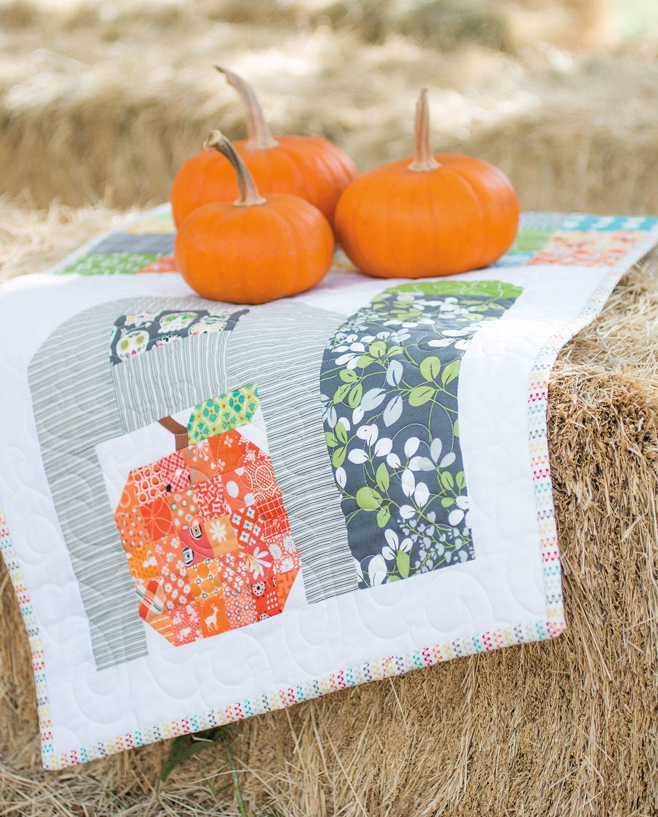 A quilted table runner of scrappy Patchwork Pumpkin blocks from Farm Girl Vintage by Lori Holt for It's Sew Emma, displayed on a hay bale with three orange pumpkins.