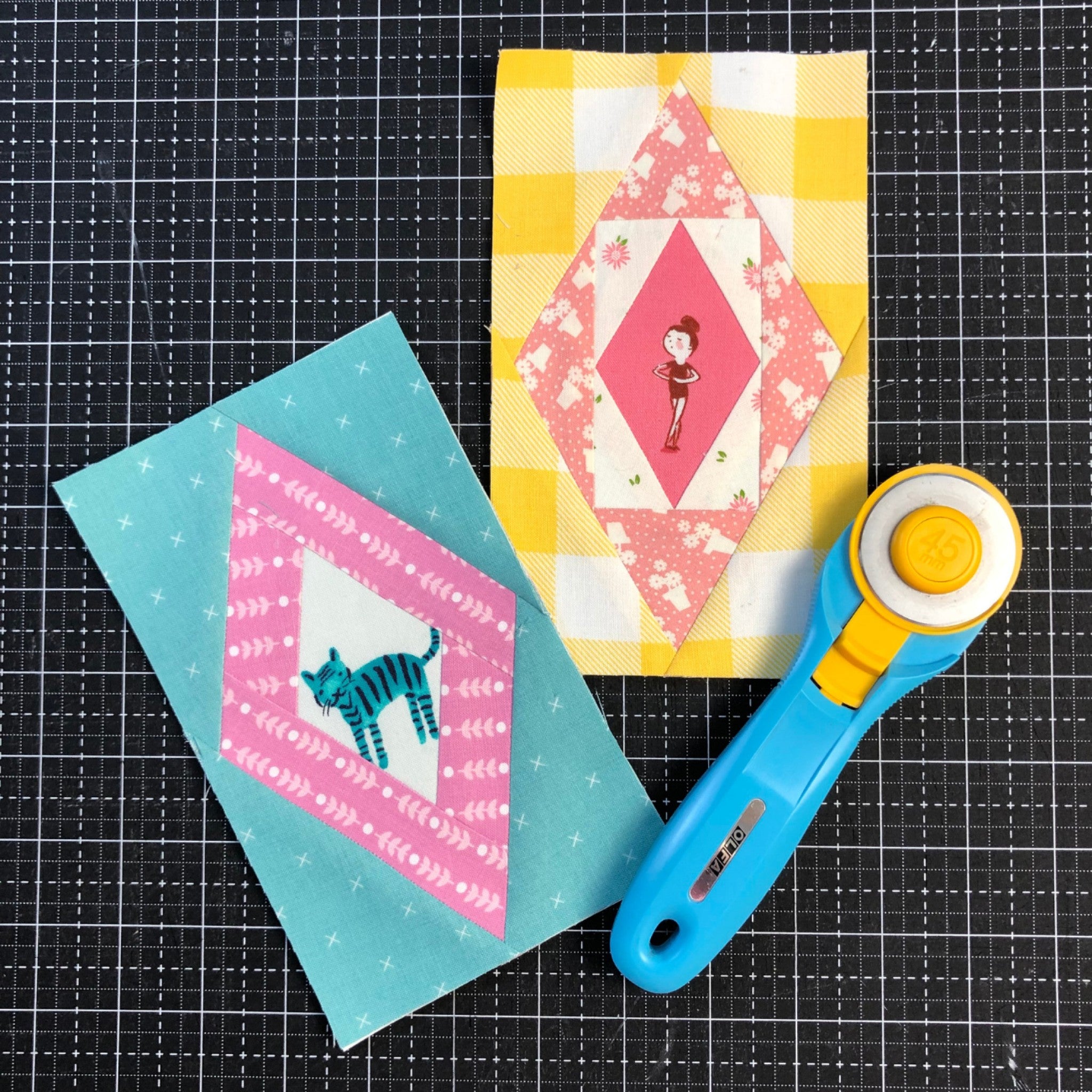Two finished Scrappy Diamond blocks from Leila Gardunia's foundation paper pad, arranged on a black cutting mat beside a blue and yellow rotary cutter. One diamond frames a fussy cut figure print in pink and cream on a yellow gingham background; the other frames a fussy cut cat print in teal and pink on an aqua background. Both show the 4 inch by 7 inch finished block size.
