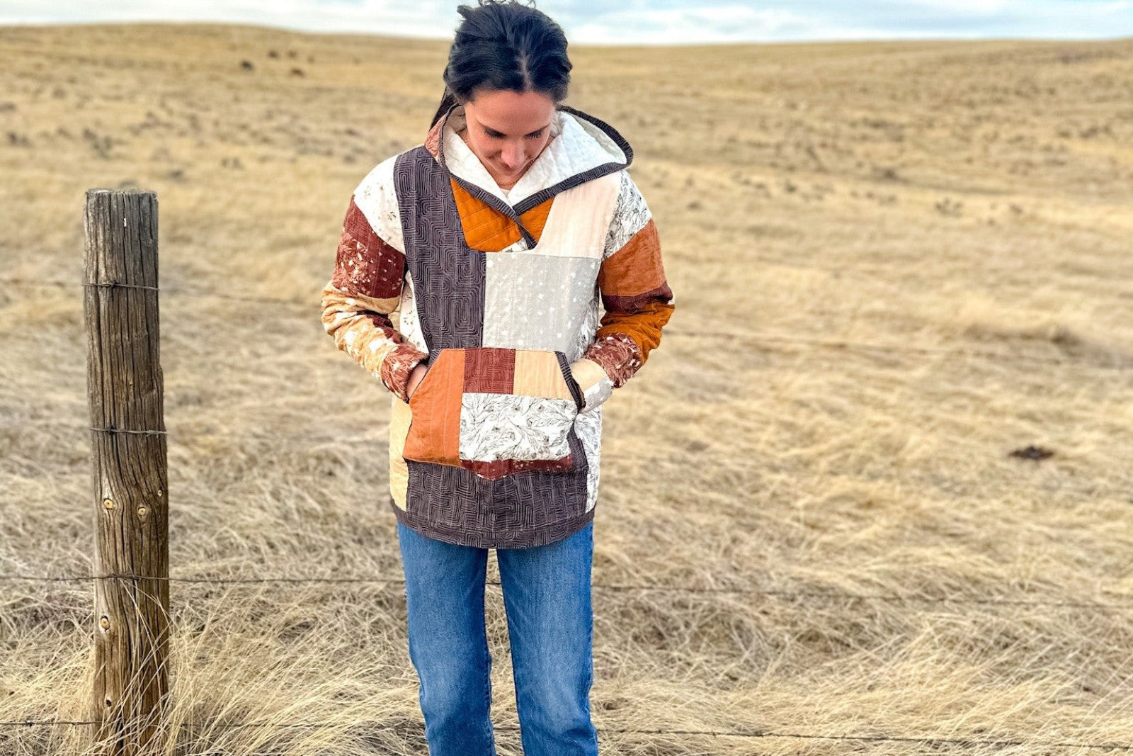 A person wearing the finished Quilted Hoodie in a rust, charcoal, and cream patchwork, hands tucked into the front kangaroo pocket, standing in a dry grass field. The pullover has a lined hood, long sleeves, and vertical channel quilting.