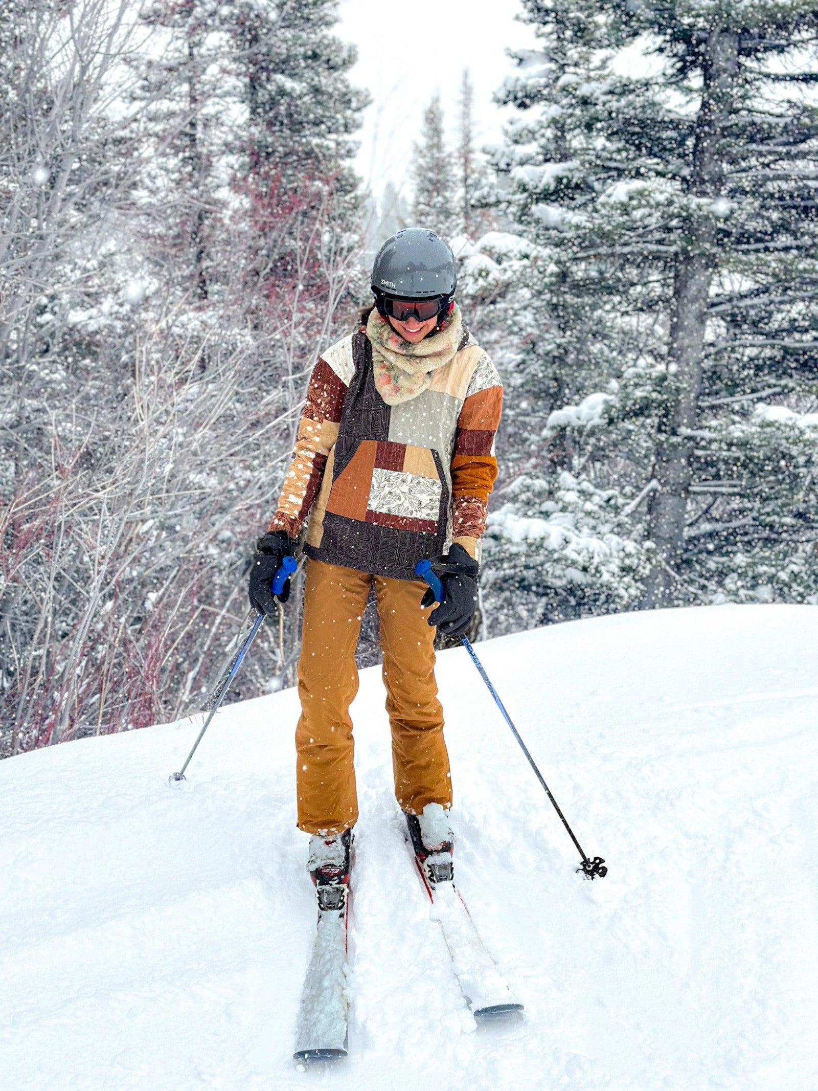 A person skiing in fresh snow while wearing the finished Quilted Hoodie in a rust, cream, and charcoal patchwork, worn as a warm outdoor layer with a lined hood and a front kangaroo pocket.