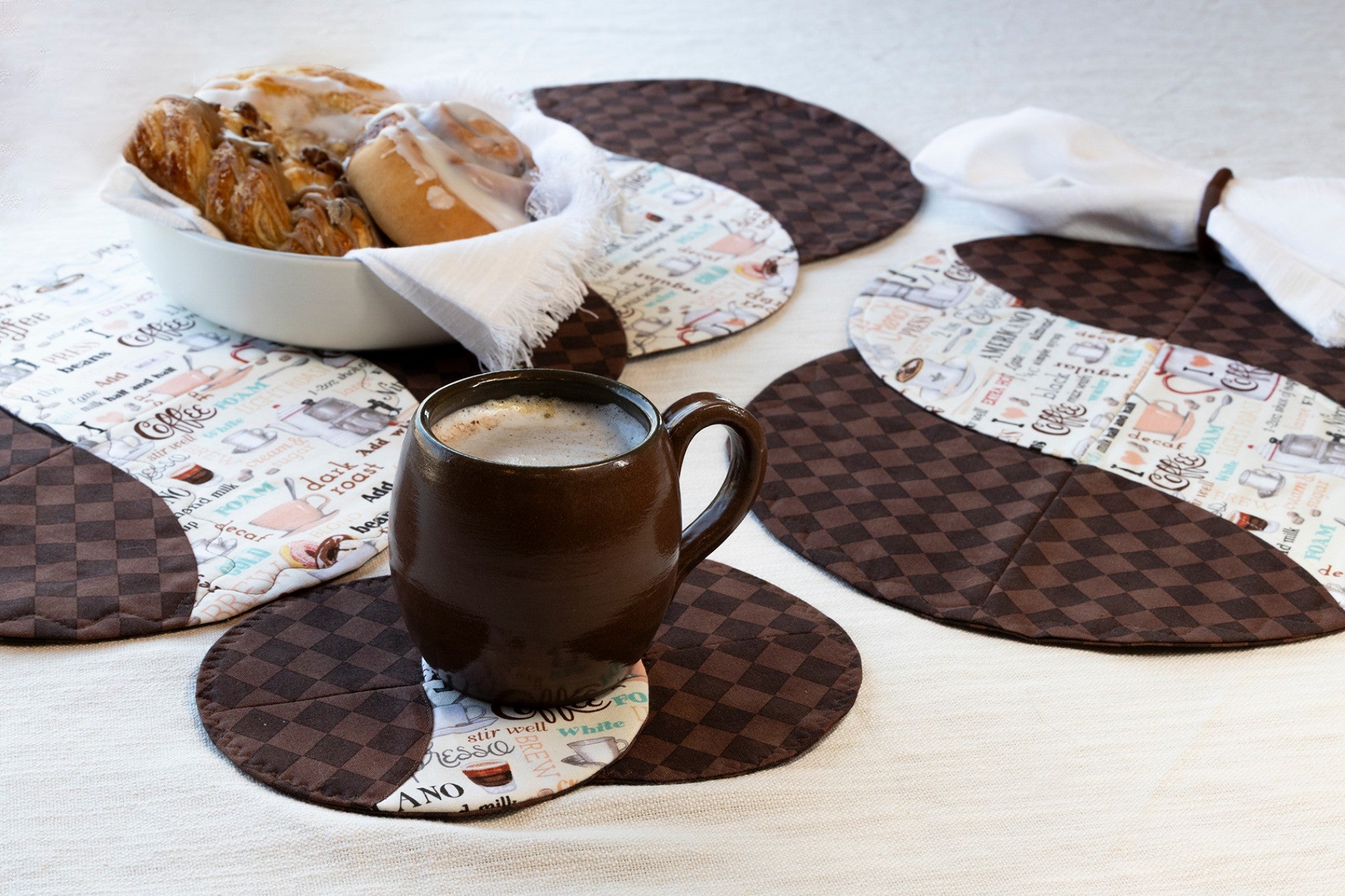 Braided Twist Junior placemats and Half Pint mug rugs in dark brown checkerboard print paired with a cream coffee-themed novelty fabric, styled on a cream linen tablecloth with a brown ceramic mug of latte and a bowl of cinnamon rolls.