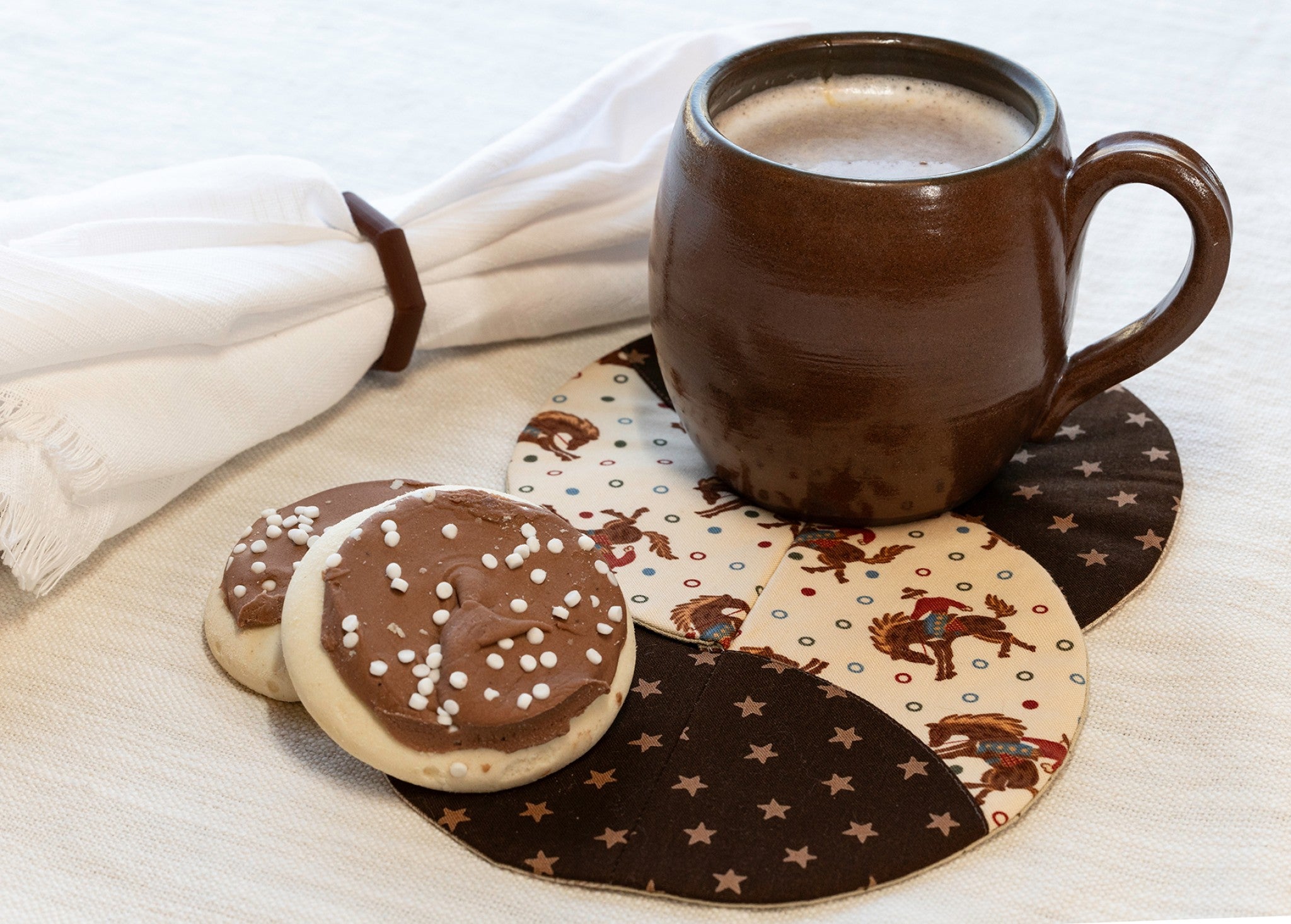 Braided Twist Half Pint mug rug in cowboy-themed quilting cotton, featuring small bucking bronco riders in brown and red on a cream background scattered with colorful polka dots, joined by a curved seam to a coordinating dark brown print covered in cream stars. Styled on a cream linen surface with a brown ceramic barrel mug of frothy latte, a folded white napkin in a brown leather napkin ring, and two chocolate-iced cookies topped with white sprinkles.