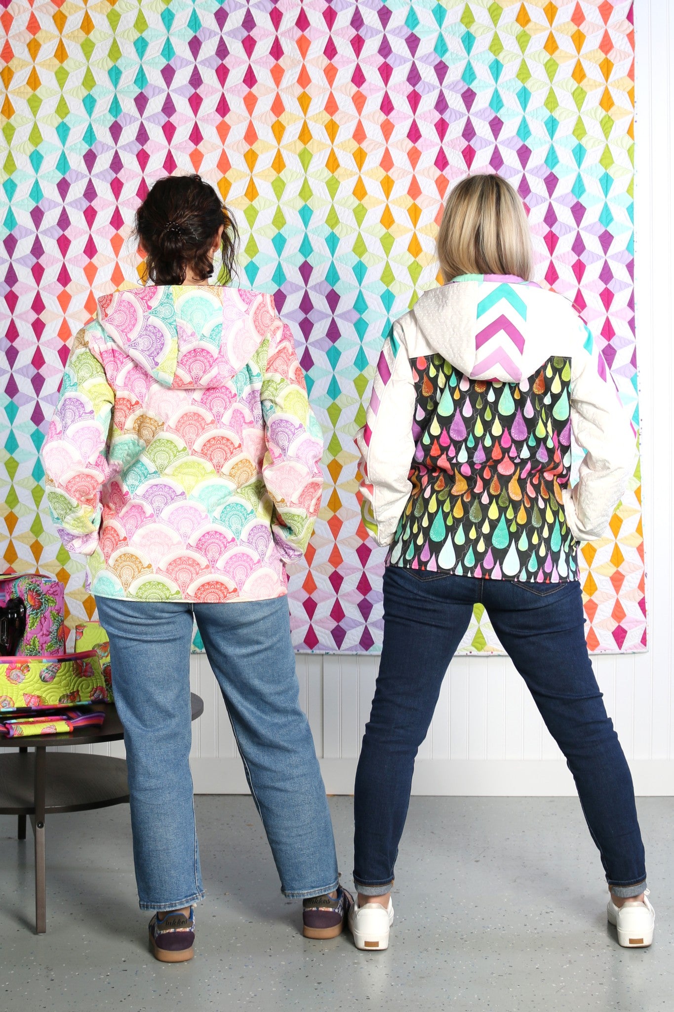 Back view of two people wearing Bella Quilt Coats, one in a pastel scallop print with the hood up and one in a raindrop print with a rainbow chevron hood down, against a colorful patterned backdrop.