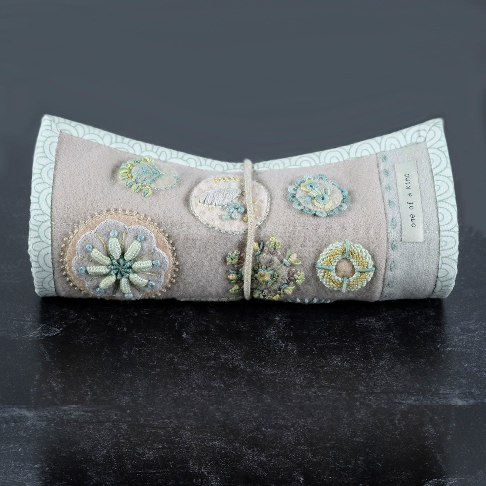 Close detail of the finished Tranquil Garden Needle Roll by Sue Spargo of Folk Art Quilts, rolled and tied with a cord, showing the tonal fog and aqua appliquéd circles worked in textural embroidery stitches with a one of a kind label.