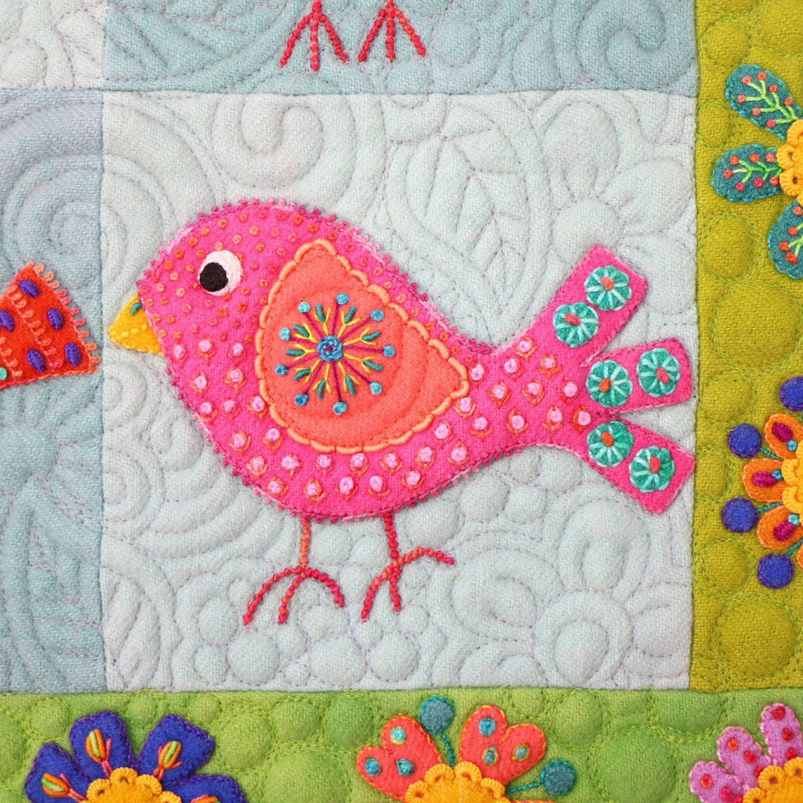 Macro close-up of a single block from Chirp by Sue Spargo of Folk Art Quilts. A pink and coral wool bird is covered in hand embroidery, including French knots and a stitched starburst on the wing, with a teal flowered tail, on a swirl-quilted pale background beside the green floral border.