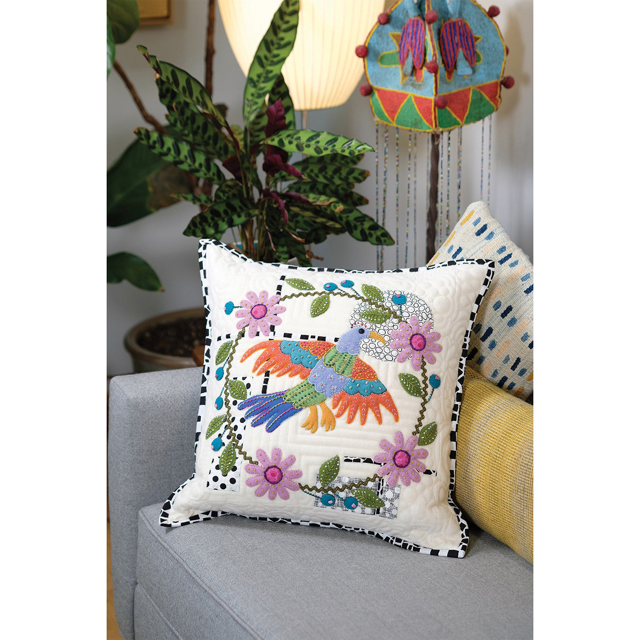 The Of a Feather Pillow, one of the book's bonus projects, a cushion with a colorful folk art bird appliqued in wool and embroidered, shown on a couch.
