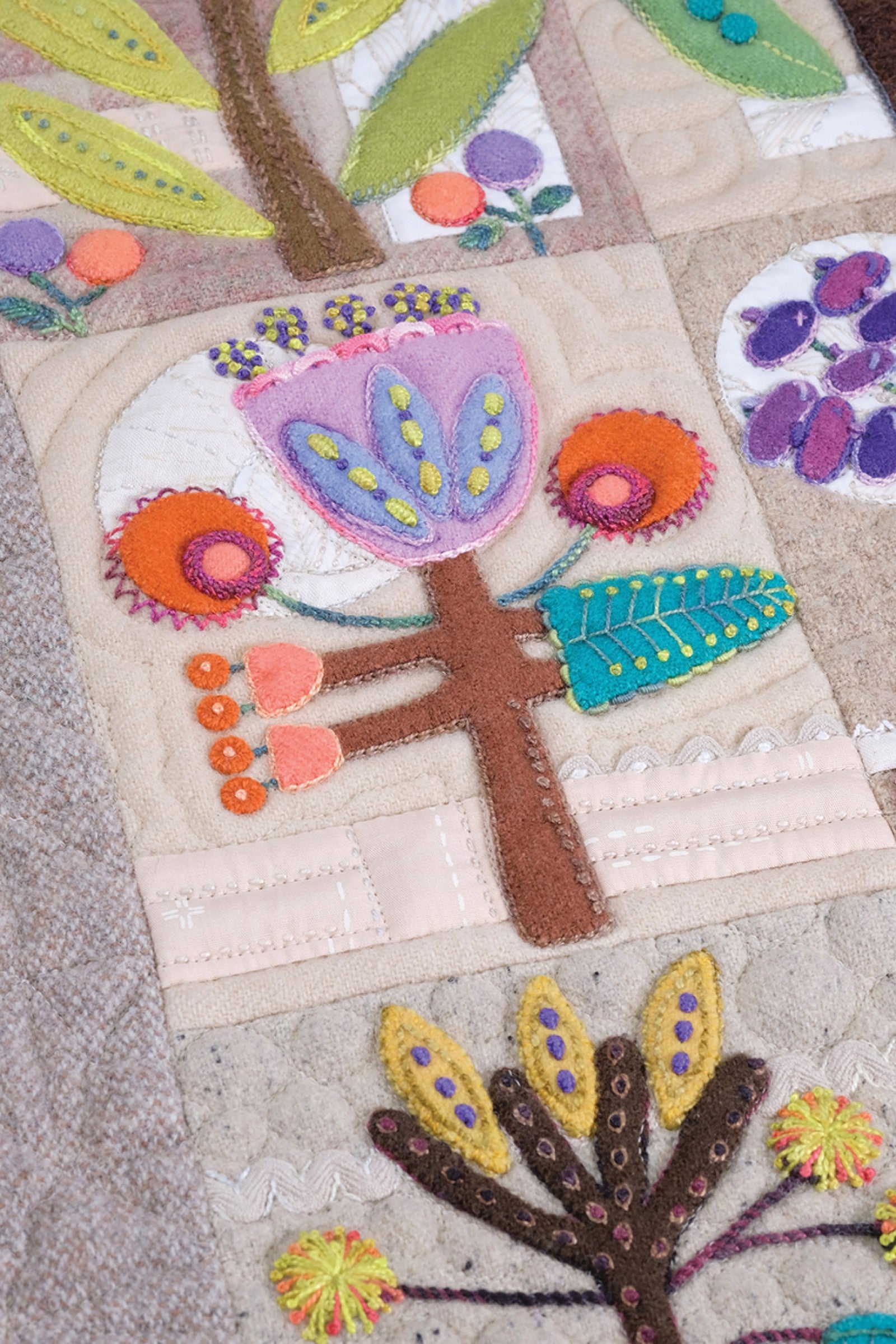 Close detail of Rooted blocks by Sue Spargo of Folk Art Quilts. A purple and pink wool bloom on a brown trunk with a teal leaf and orange berries, above a yellow-petal flower, in layered wool and cotton applique with hand embroidery.
