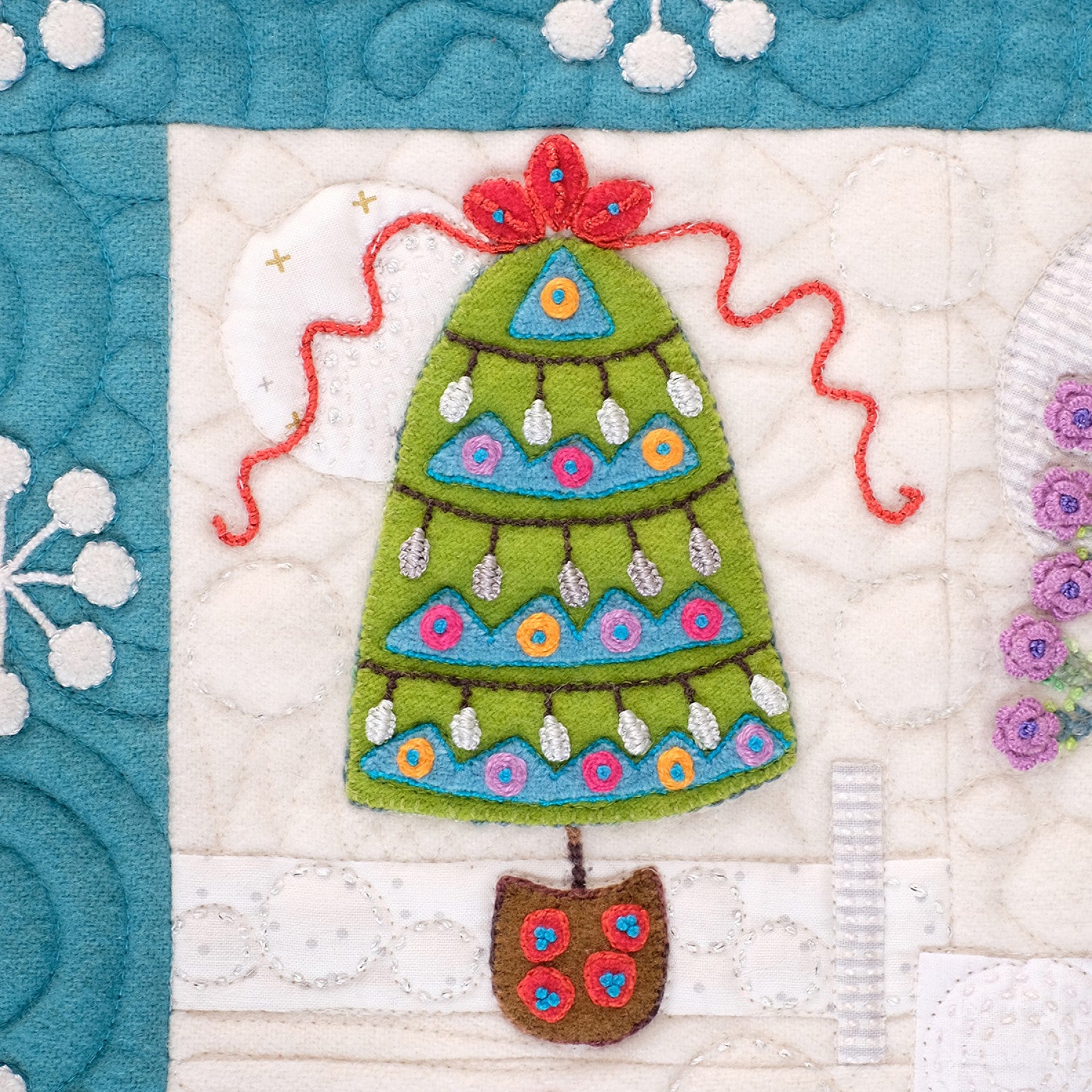 Close detail of a Tinsel block by Sue Spargo of Folk Art Quilts. A green wool Christmas tree with embroidered lights, flowers, and a red topper sits above a small pot, on a snowflake-quilted white background with a blue border edge.