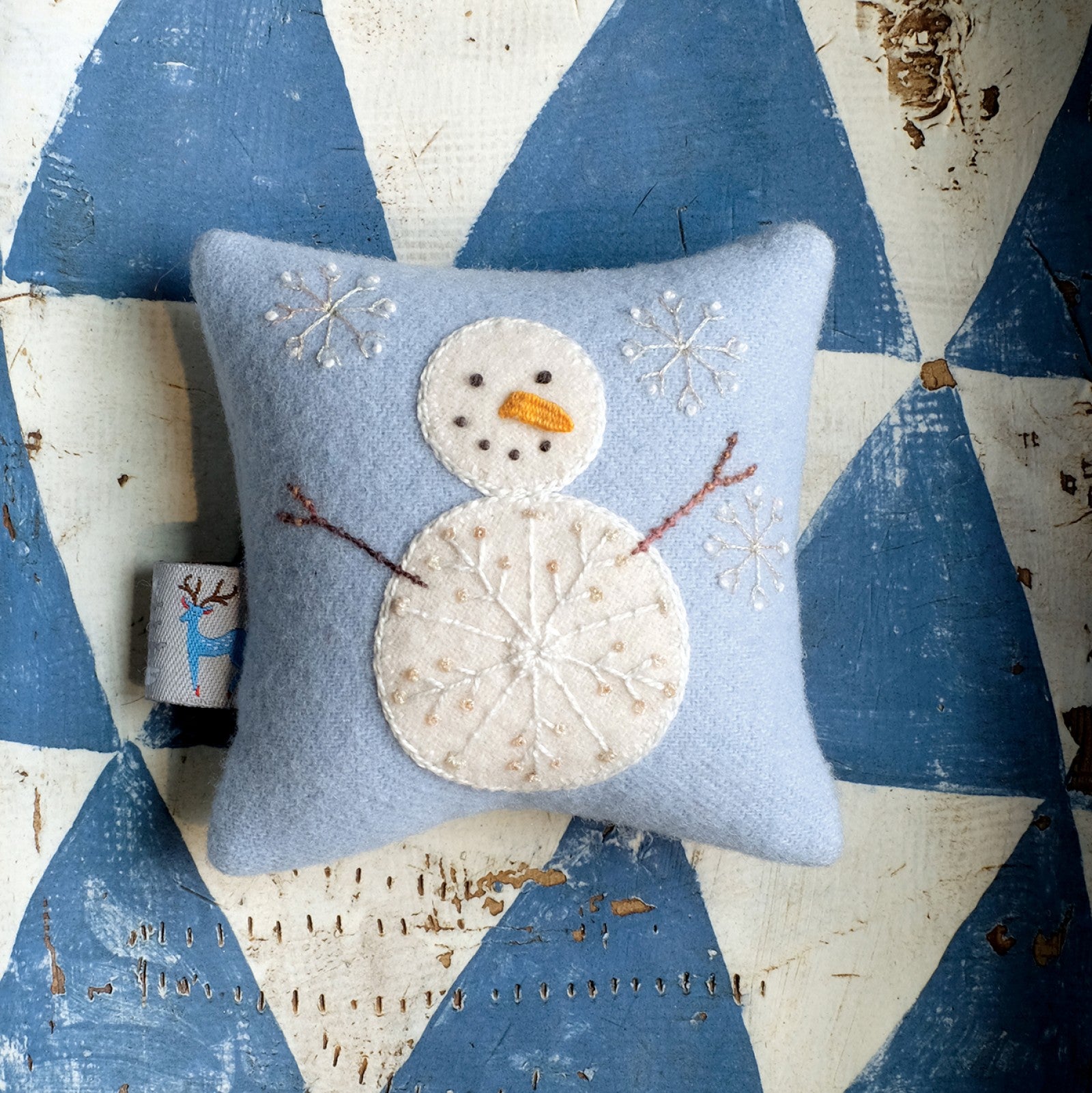 The bonus Snowman pincushion from Tinsel by Sue Spargo of Folk Art Quilts, a soft blue wool pincushion with an appliqued snowman and embroidered snowflakes, set on a blue and white background.