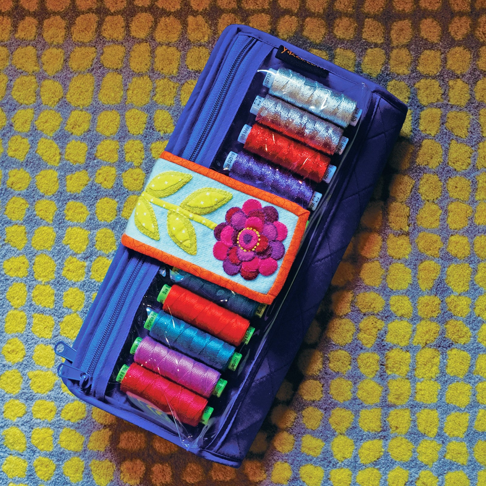 The bonus Tinsel Workbox from Sue Spargo of Folk Art Quilts, a blue and purple clutch-style organizer holding embroidery thread spools, with an appliqued flower motif on the front.