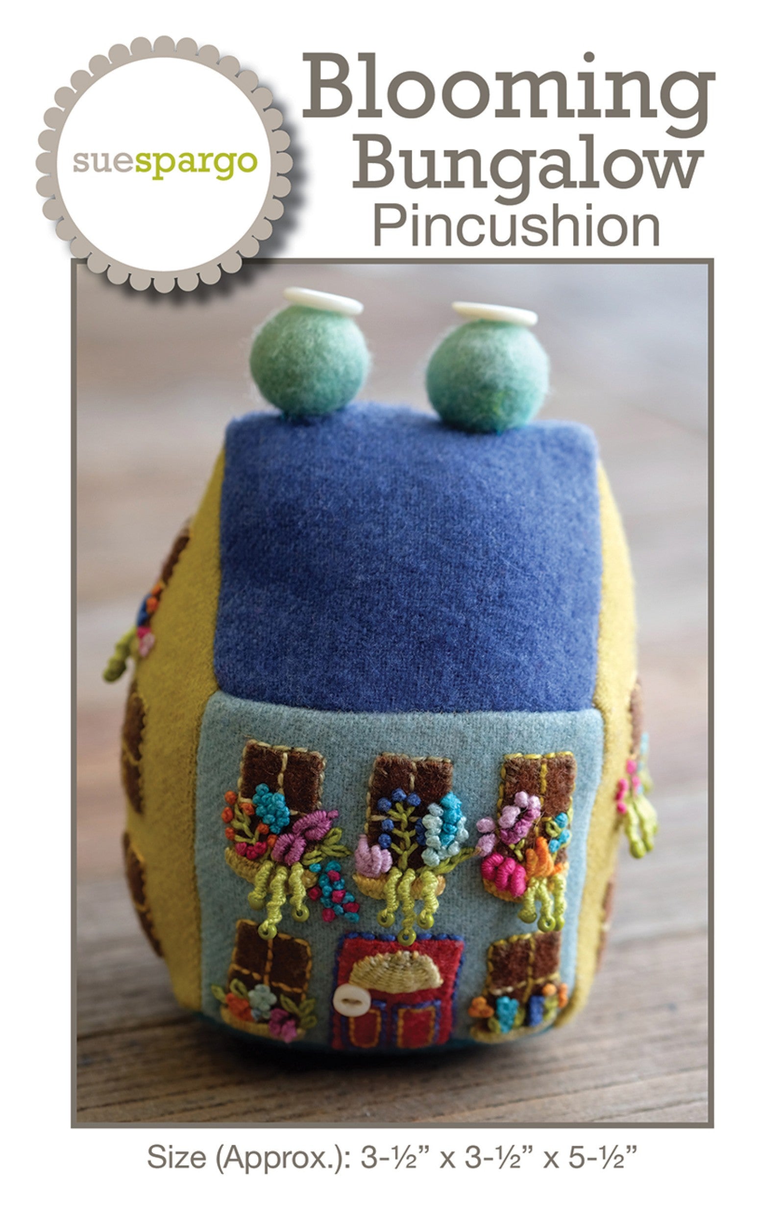 Blooming Bungalow Pincushion sewing pattern by Sue Spargo of Folk Art Quilts. Front cover shows the finished three-dimensional house pincushion in yellow wool with a blue roof and two button-topped chimneys, a sage front with appliquéd windows and a red door, and window boxes filled with embroidered flowers. Size about 3-1/2 by 3-1/2 by 5-1/2 inches shown.