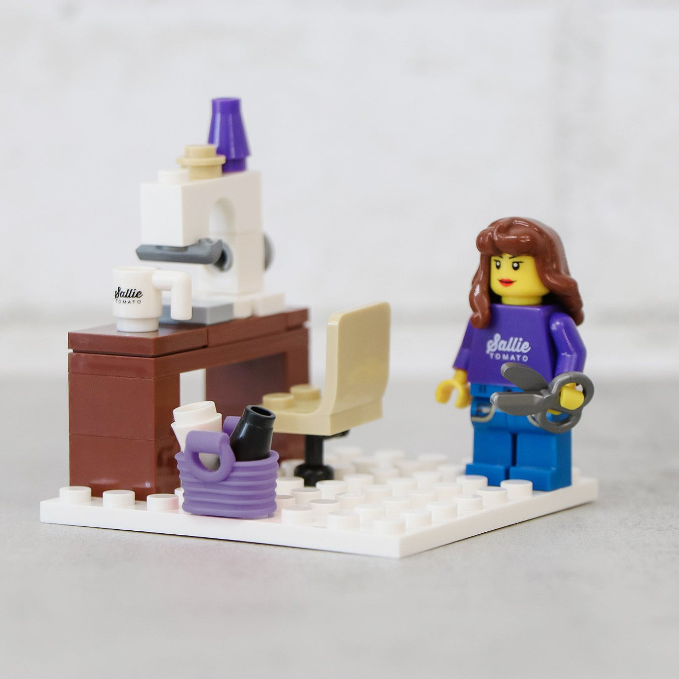 Finished Sallie Tomato Sewist Building Block Set: a built sewing-room scene with a white sewing machine on a brown work table, a swivel chair, thread and scissors, and a sewist figurine in a purple Sallie Tomato shirt, on a white base.
