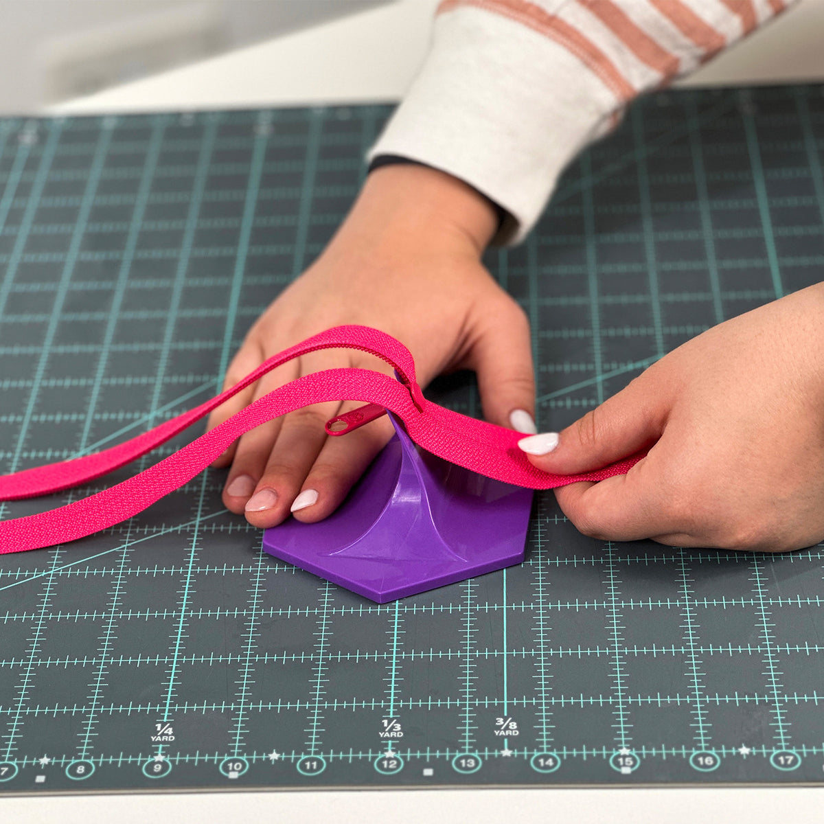 Hands threading pink zipper tape into a pull that sits in the purple Handy Zipper Jig on a green cutting mat.
