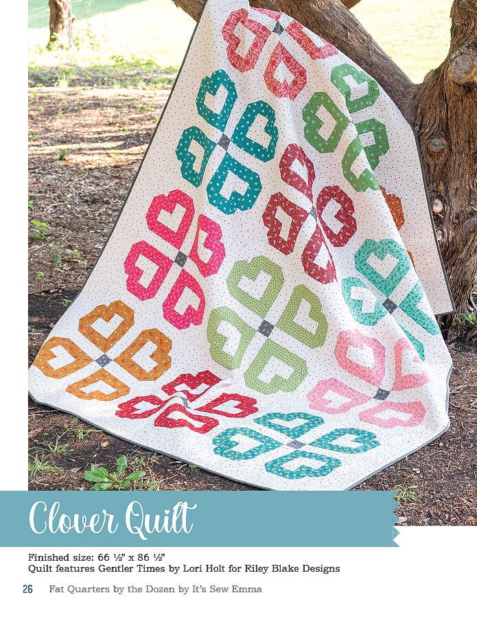 The Clover quilt, 66.5 x 86.5 inches, four-leaf clover blocks in rainbow prints on white, draped by a tree.