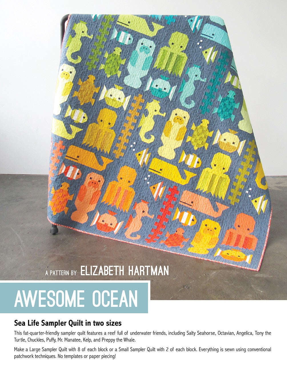 Awesome Ocean quilt pattern cover by Elizabeth Hartman. Large 71 by 83 inch quilt propped on a stand shows nine patchwork sea creature blocks including seahorse, octopus, jellyfish, turtle, clownfish, pufferfish, manatee, kelp, and whale arranged in a rainbow gradient from cool greens and teals at top to warm oranges and corals at bottom on a dark blue-gray background.