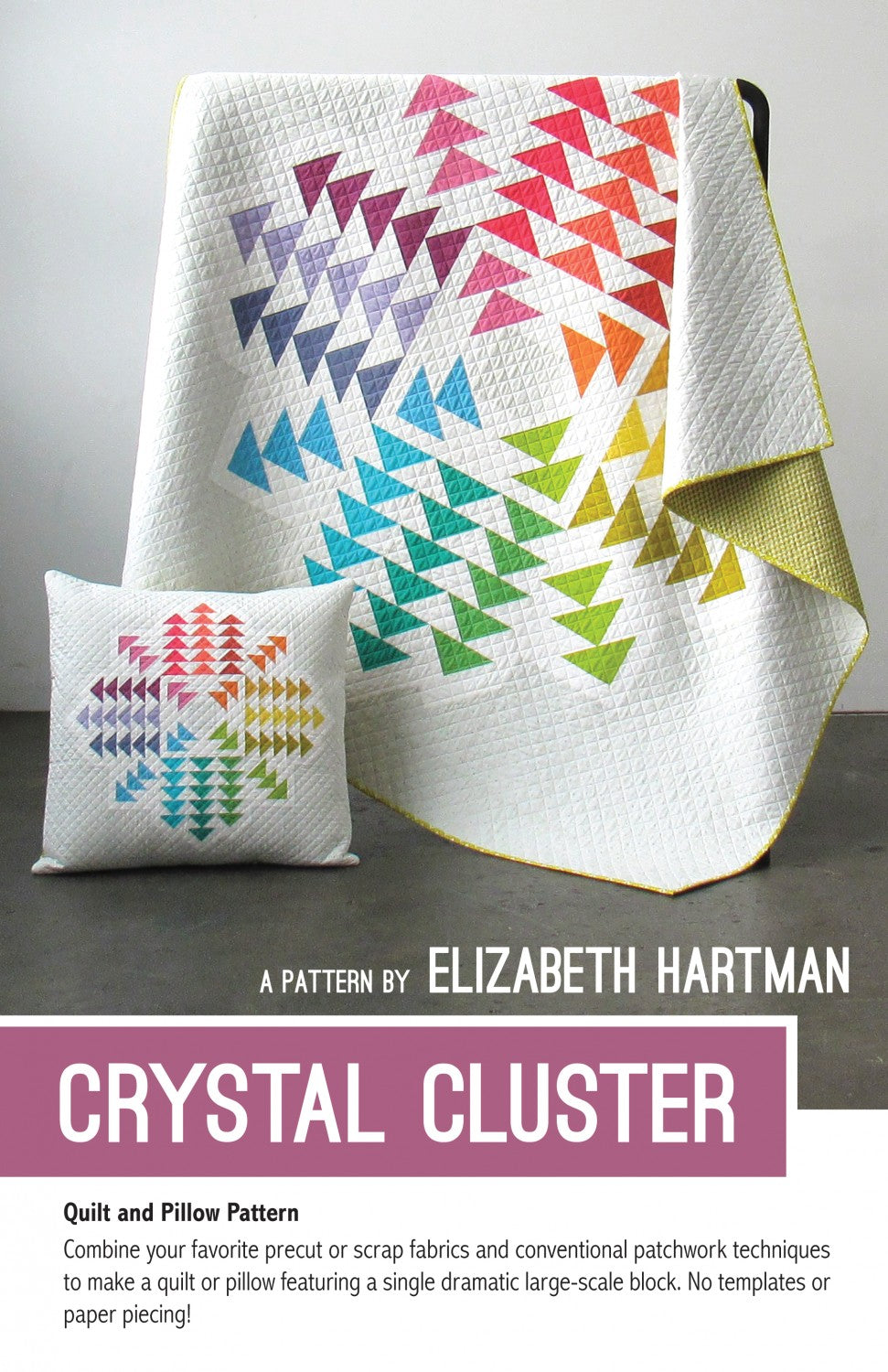 Crystal Cluster quilt pattern cover by Elizabeth Hartman. The 70 by 70 inch lap quilt leans on a stand showing a large symmetrical crystal starburst made from flying geese units in 16 rainbow colors -- pink, red, orange, yellow, green, teal, blue, and lavender -- on a white background. A 24 by 24 inch pillow in the foreground shows the same design in miniature.