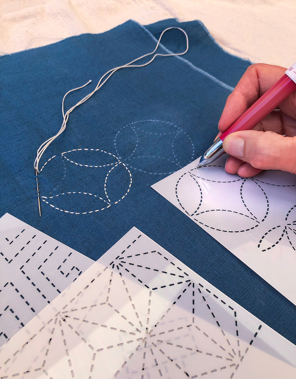 A person traces a sashiko design through the dashed slots of a translucent Sylvia Pippen stencil with a pink marking pencil on blue cloth. Beside it, white sashiko thread and a needle follow the dashed marks of an overlapping-circle design, and two more stencils lie in front.