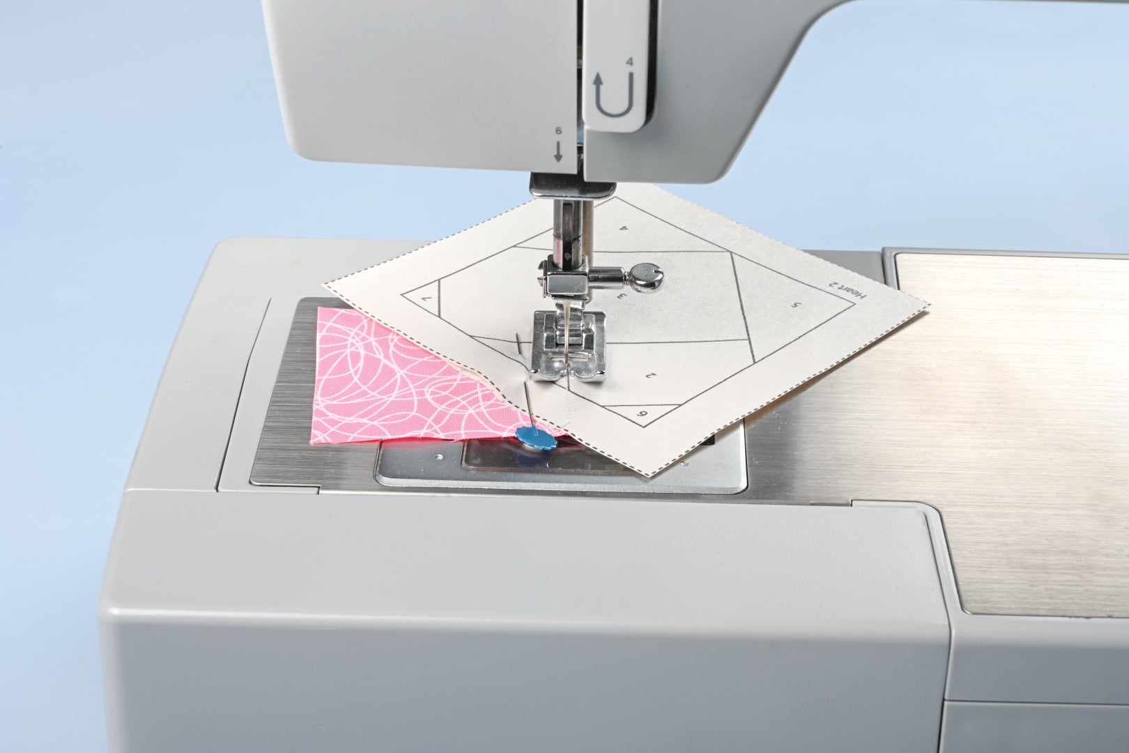 A foundation pattern printed on Carol Doak's Foundation Paper from C&T Publishing feeding under the presser foot of a sewing machine. Pink scribble-print fabric is held to the underside by a blue-headed pin, and the printed lines and numbered sections show through the sheet.