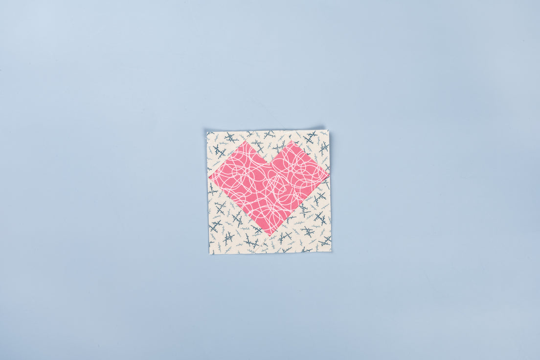 A finished heart quilt block pieced on Carol Doak's Foundation Paper from C&T Publishing and shown with the paper torn away. A pink scribble-print heart sits on a cream background printed with small navy and grey crosses.