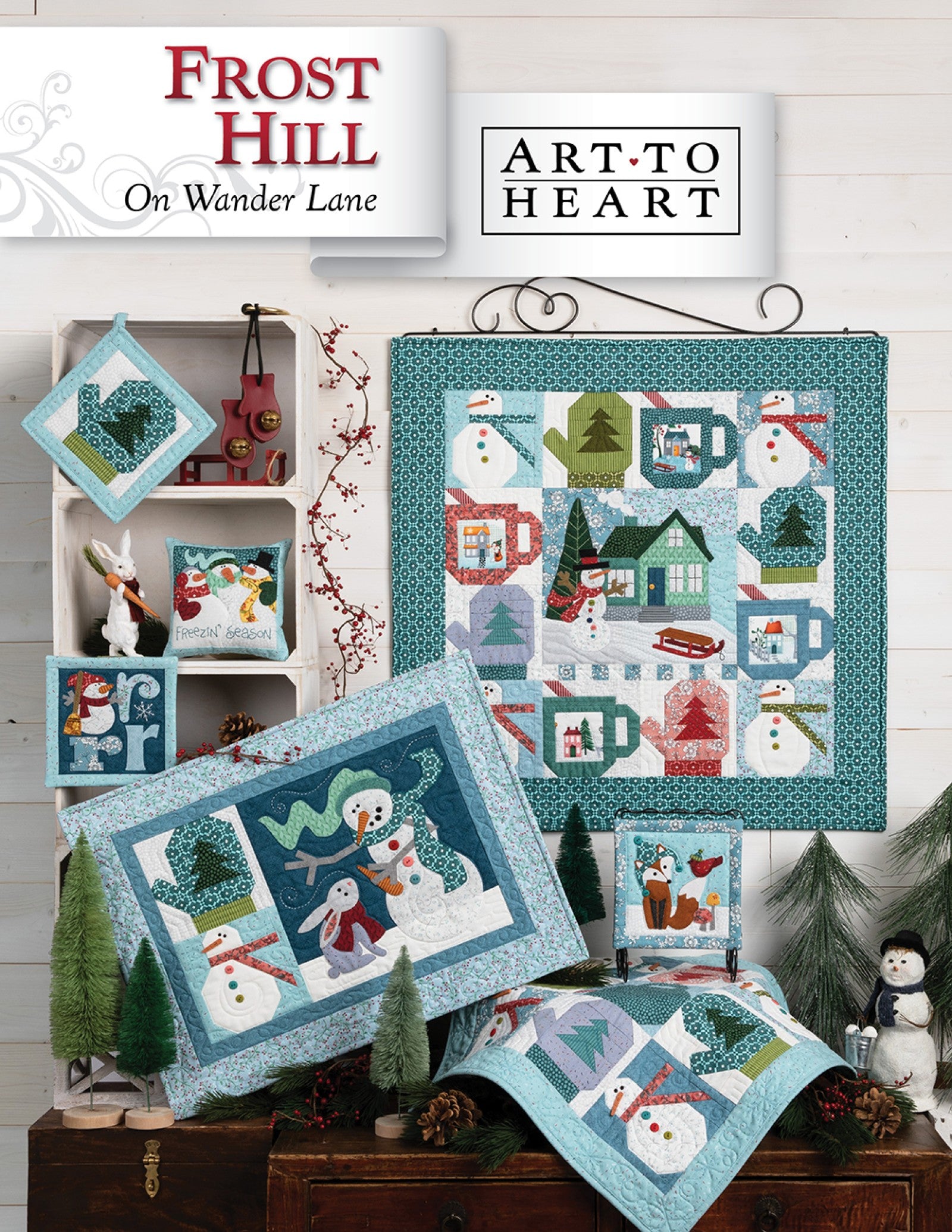 Front cover of the Frost Hill on Wander Lane quilt pattern by Nancy Halvorsen, Art to Heart, showing the snowy winter village appliqué block framed in patchwork with a snowman pillow and cool blue bonus projects.