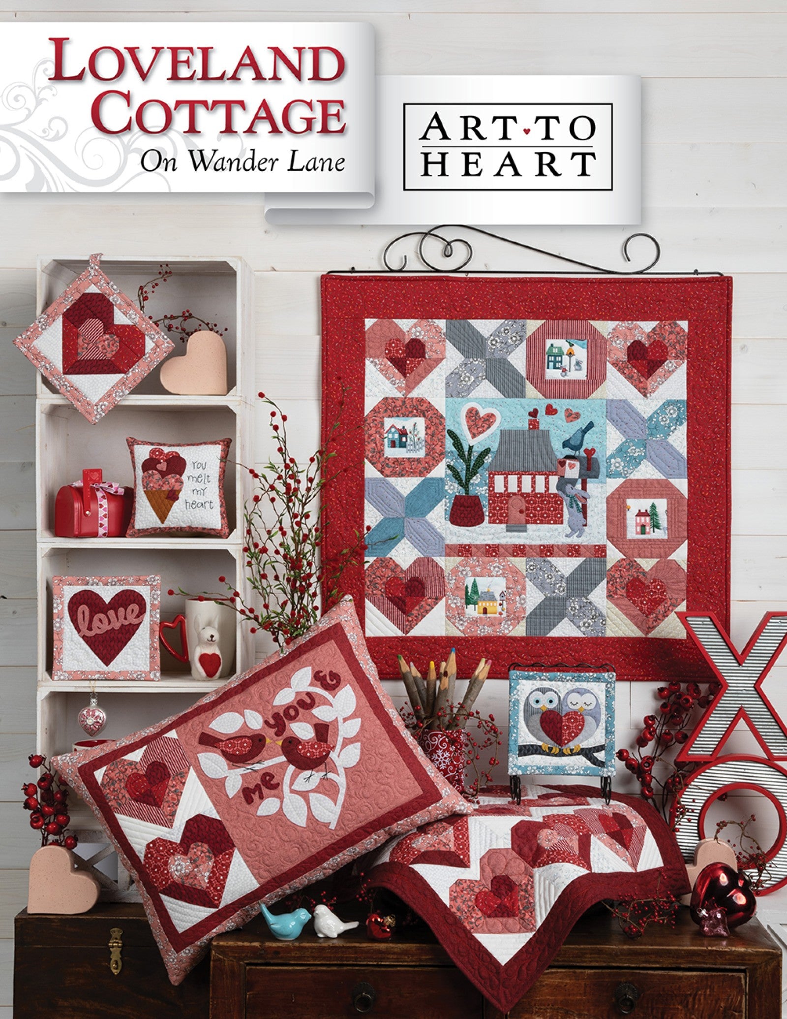 Front cover of the Loveland Cottage on Wander Lane quilt pattern by Nancy Halvorsen, Art to Heart, showing the heart and cottage appliqué block framed in patchwork with Valentine pillows and mini quilts in reds and pinks.