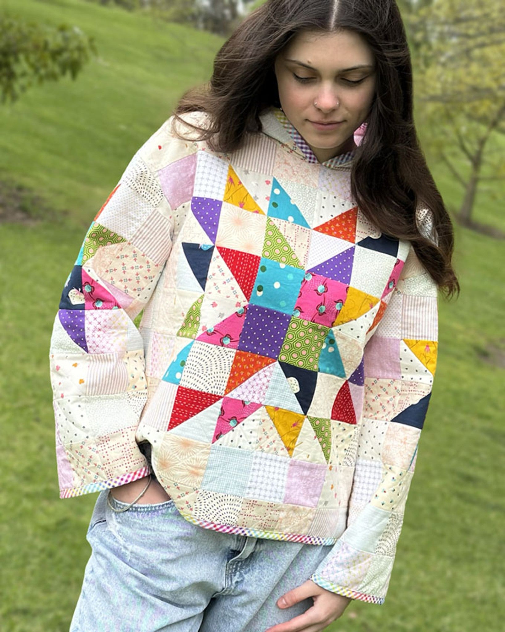 Lexi Hoodie by Melanie McNeice for Melly & Me, front view. A person with long dark hair wears the patchwork hoodie with the hood down, showing the bright pieced star on a scrappy cream and pastel background and rainbow gingham trim.