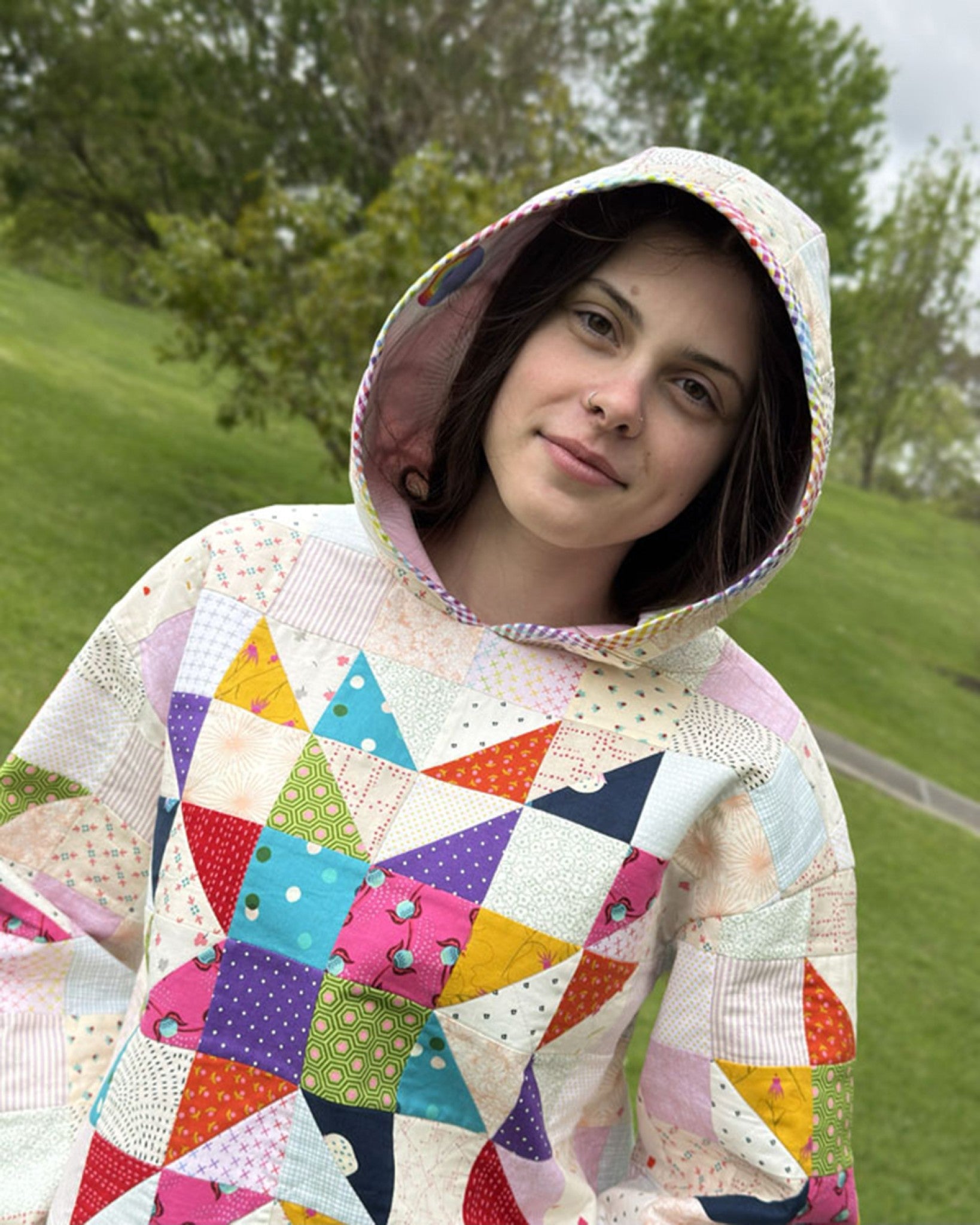 Lexi Hoodie by Melanie McNeice for Melly & Me. A person wears the patchwork hoodie with the hood up, rainbow gingham binding framing the hood opening, and the bright pieced star and scrappy pastel patchwork visible on the body.